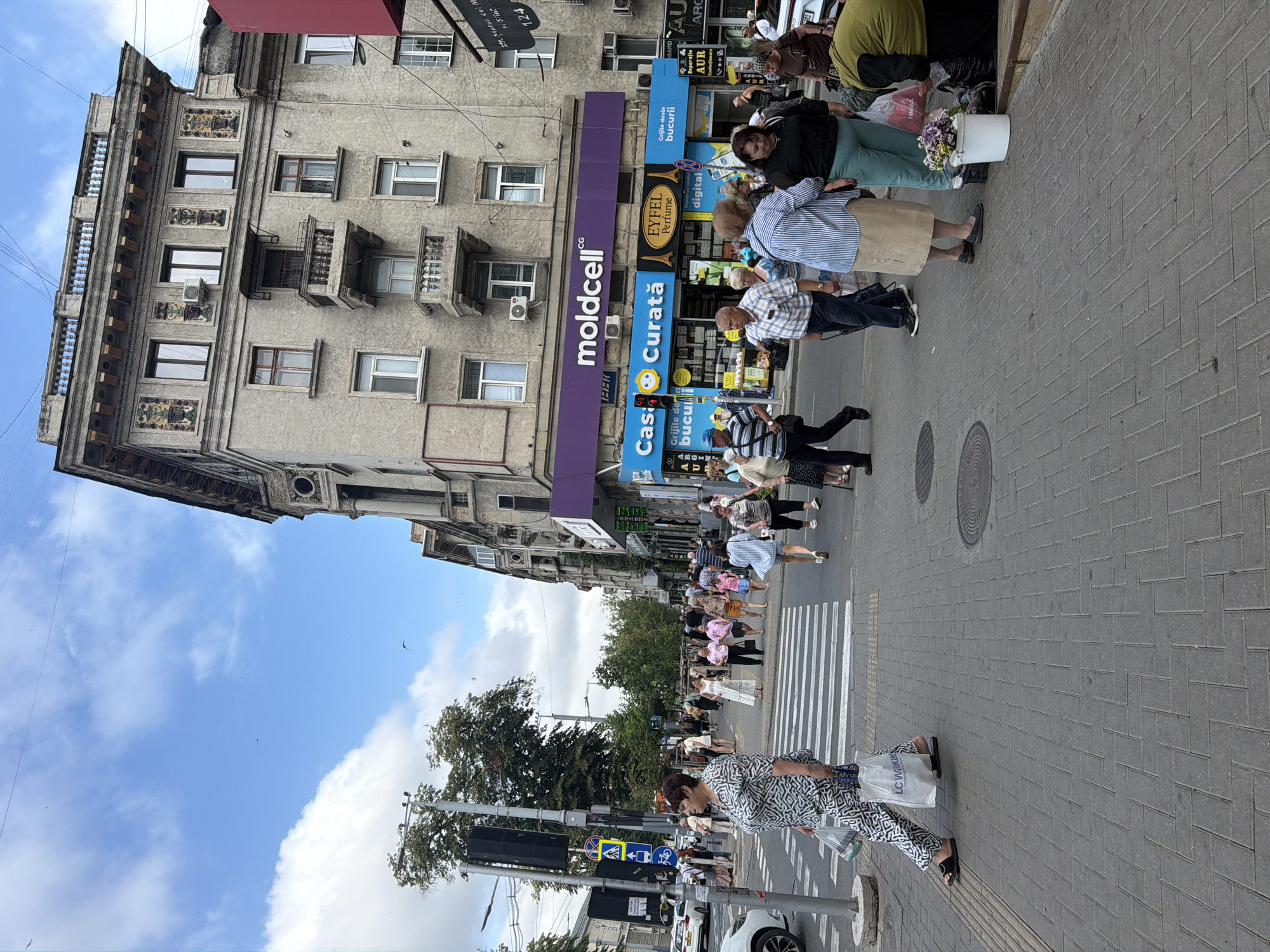 The image depicts a busy street scene in Chișinău, Moldova, likely on a warm, cloudy day. In the foreground, a woman with dark, short hair, wearing a black and white patterned dress and black sandals, walks across a pedestrian crossing, carrying two shopping bags, one white with "LC Waikiki" printed on it. The street is paved with grey interlocking bricks. Behind her, several other pedestrians are crossing the street or walking on the sidewalk.
The buildings lining the street are a mix of architectural styles. On the left, a tall, grey apartment building with ornate architectural details and balconies dominates the view. Below it, a traffic light and street signs are visible. On the right, a modern storefront with a purple awning displays the "moldcell" logo, and below it, a bright blue storefront features signs for "Grijile devin bucurii" (Worries become joys), "EYFEL Perfume," and other businesses. The street is lined with trees and electrical wires are visible against the sky. In the distance, more buildings and people suggest an urban environment.