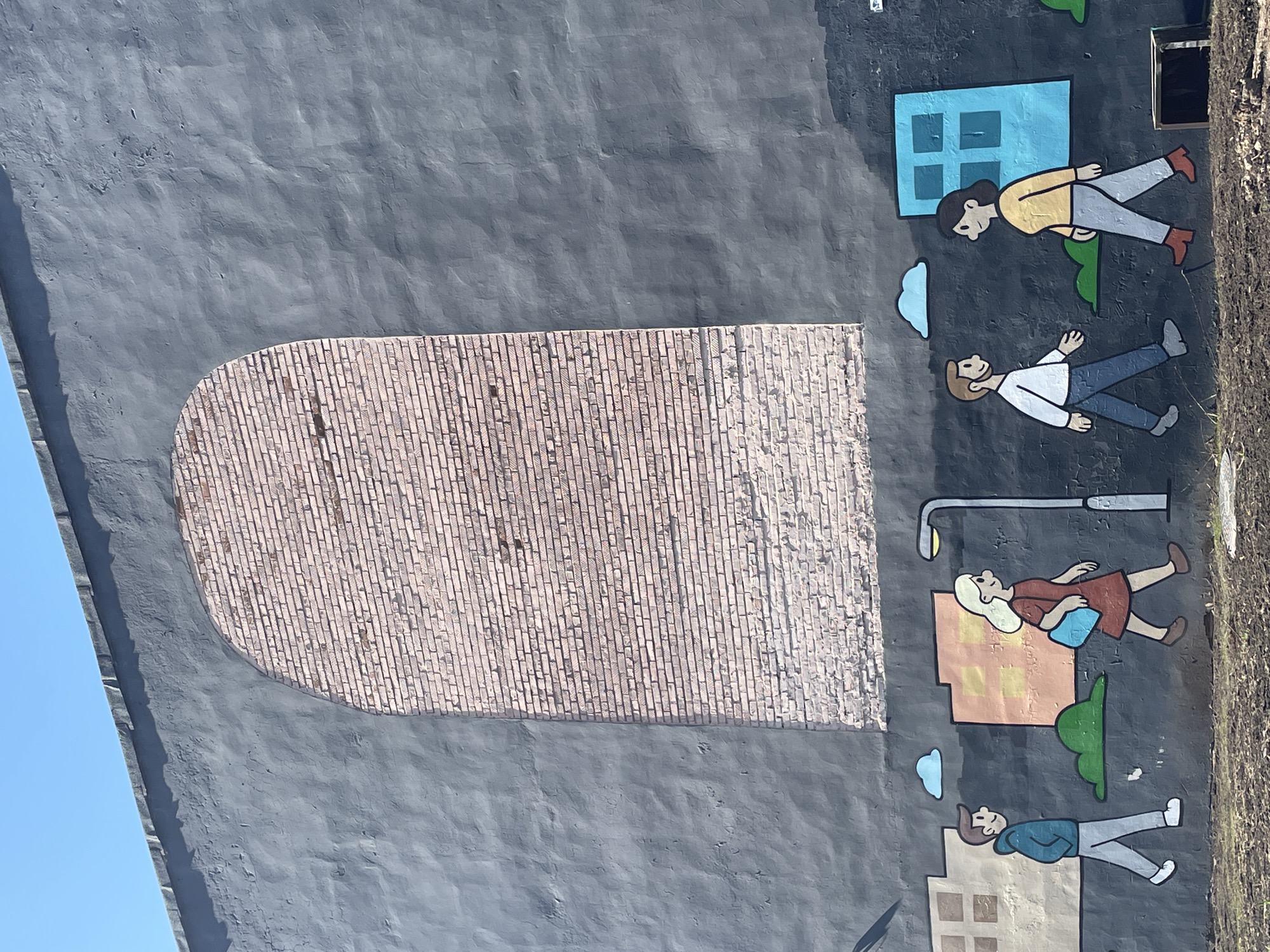 The image depicts a mural on the side of a building, presumably located in Izhevsk, Russia. The background is painted in a muted gray color and has a roughly textured appearance. A large, arched section filled with a brick pattern dominates the central part of the mural. To the right of this brick area, cartoon-style figures of people are painted with simplified features and flat colors. Among them are stylized buildings, a street lamp, and puffy clouds, adding to the whimsical nature of the mural.