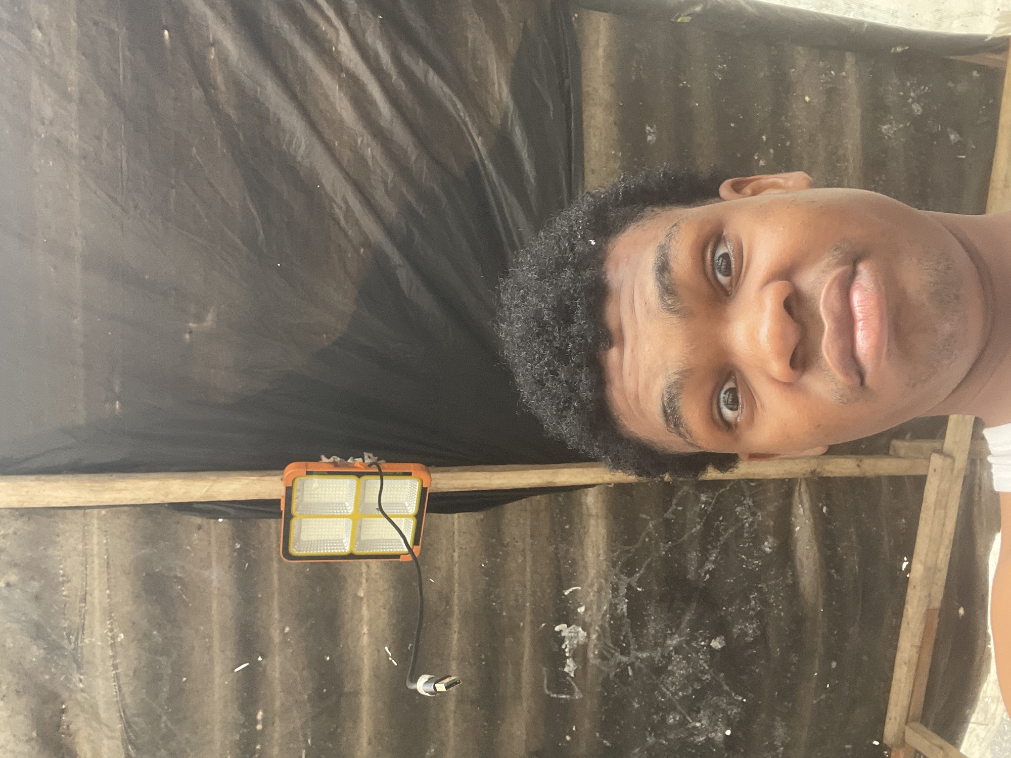 A medium shot captures a young Black male with curly hair looking directly at the camera with a surprised expression, his eyes wide open and his mouth slightly agape. He is wearing a white tank top. The background is dark and textured, possibly a corrugated metal roof covered with a black tarp, with wooden supports visible. To the left, an orange and white LED floodlight is mounted on a wooden beam, with a black cable attached. The environment appears to be an outdoor, possibly unfinished, structure. The location is identified as Ilado, Nigeria.