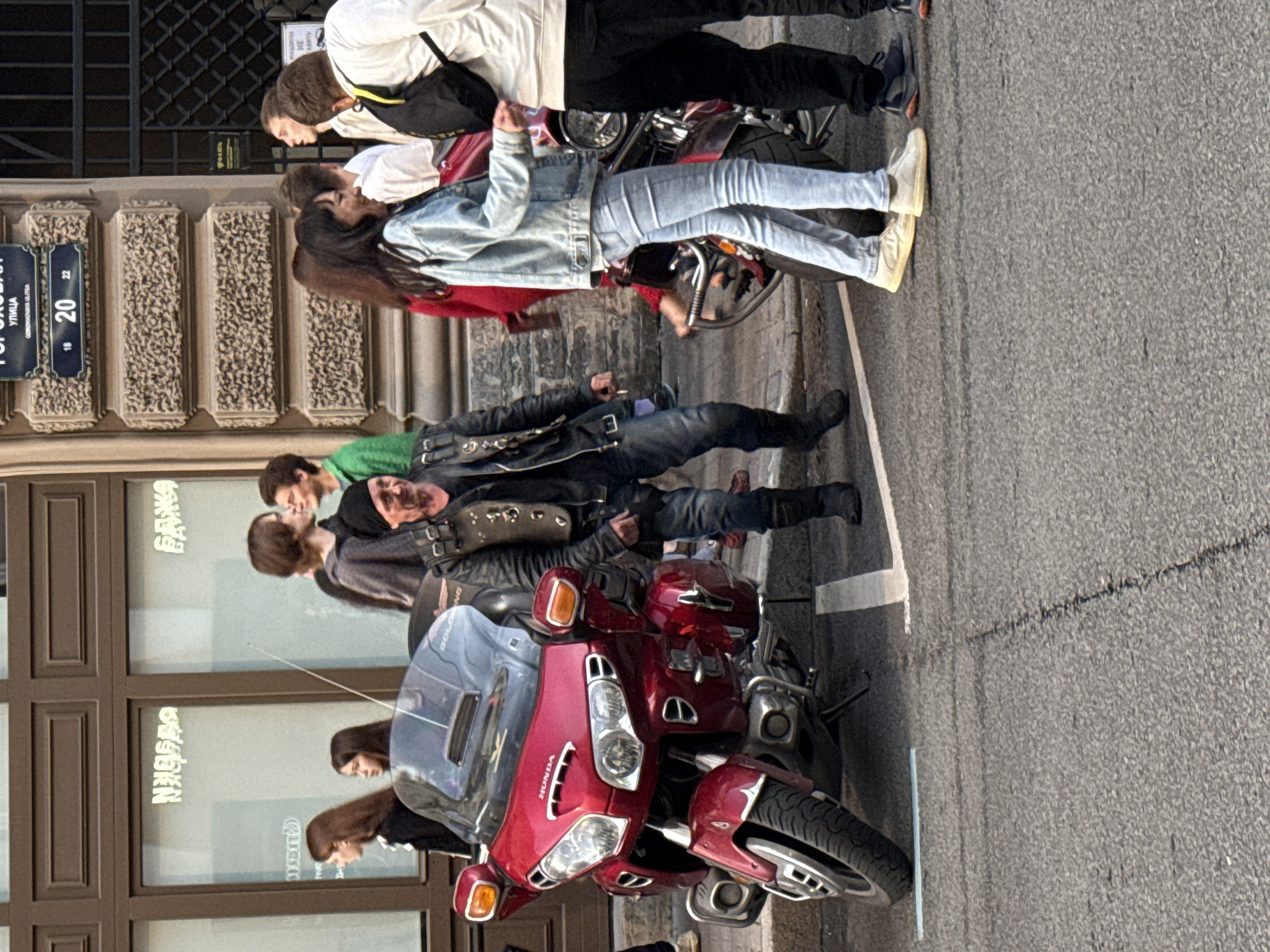 The image depicts a street scene in Saint Petersburg, Russia, likely on or near Gorokhovaya Street, as indicated by the street sign showing "ГОРОХОВАЯ УЛИЦА" and numbers "18 20 22". In the foreground, a large red Honda motorcycle is parked on the asphalt, distinguished by its spacious fairing and windshield. A group of people are gathered around it.
A man in a black leather jacket, black t-shirt, black beanie, and dark jeans is the central figure, standing to the right of the motorcycle. He is looking towards the right side of the frame. To his right, a woman in a light blue denim jacket and light blue jeans, with dark hair, is facing away from the camera, talking to a person partially visible to her right. A man in a white hooded sweatshirt with a black backpack worn across his chest is standing behind her, facing left.
To the left of the motorcycle, two women with long brown hair are standing near a building entrance. One is wearing a black jacket, and the other is wearing a light-colored top. Further to the left, a man in a grey hooded sweatshirt is partially visible, looking towards the right. In the background, several other individuals are present, some of whom appear to be engaged in conversation. The building in the background features large windows and textured stone detailing, typical of St. Petersburg's architecture. The overall atmosphere appears to be a casual street gathering.