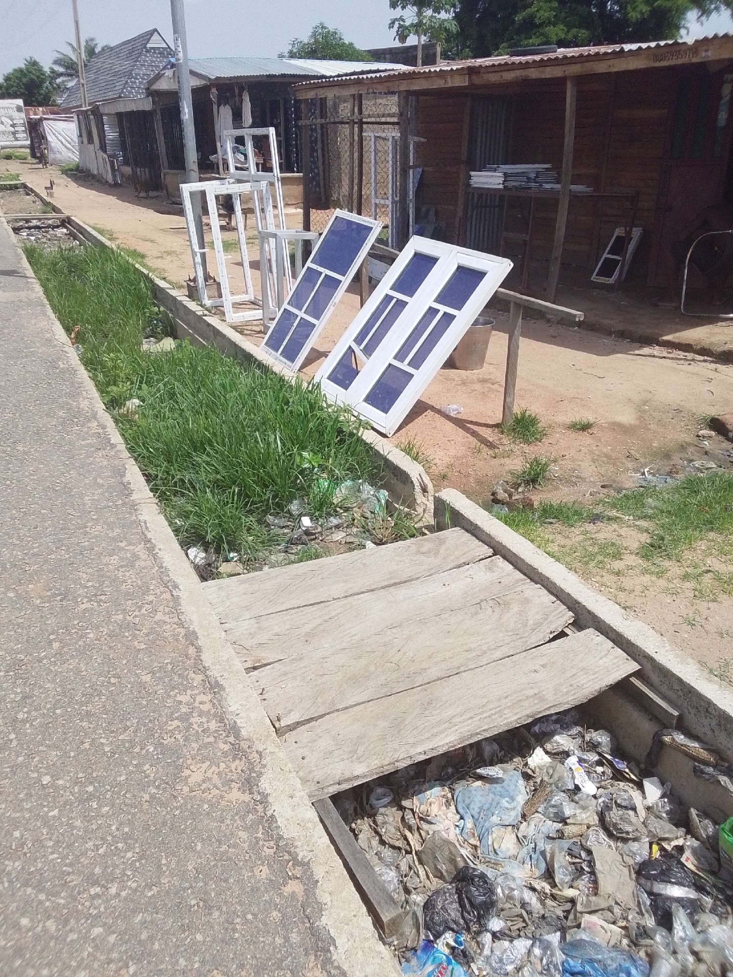The image depicts an outdoor scene in what appears to be a commercial or workshop area. In the foreground, a concrete curb lines a ditch filled with lush green grass and a significant amount of plastic waste, including bags, bottles, and other debris. A rudimentary wooden bridge spans across the ditch, made of three planks.
Further back, the scene opens up to a sandy or dirt ground. Several white window frames, some with blue tinted glass, are displayed standing upright and leaning against supports, suggesting a window manufacturing or sales business. Beyond these, a series of structures are visible. The one on the left has a dark-colored roof with a textured pattern and a light-colored facade. Adjacent to it is a longer, open-fronted structure with a corrugated metal roof and wooden walls, resembling a workshop or market stall. Inside this structure, more window frames are stacked on shelves, and a ladder leans against a wall. Another building to the right appears to be made of stone or concrete blocks with a similar corrugated metal roof.
The overall setting is somewhat cluttered and appears to be a street-side business in a developing area. The time of day seems to be daytime, with clear, bright lighting casting shadows, indicating sunny weather. There are no people visible in the image. A partially obscured phone number or identifier, "01039359712," is visible on the corrugated roof of one of the wooden structures. The environment suggests a warm climate.