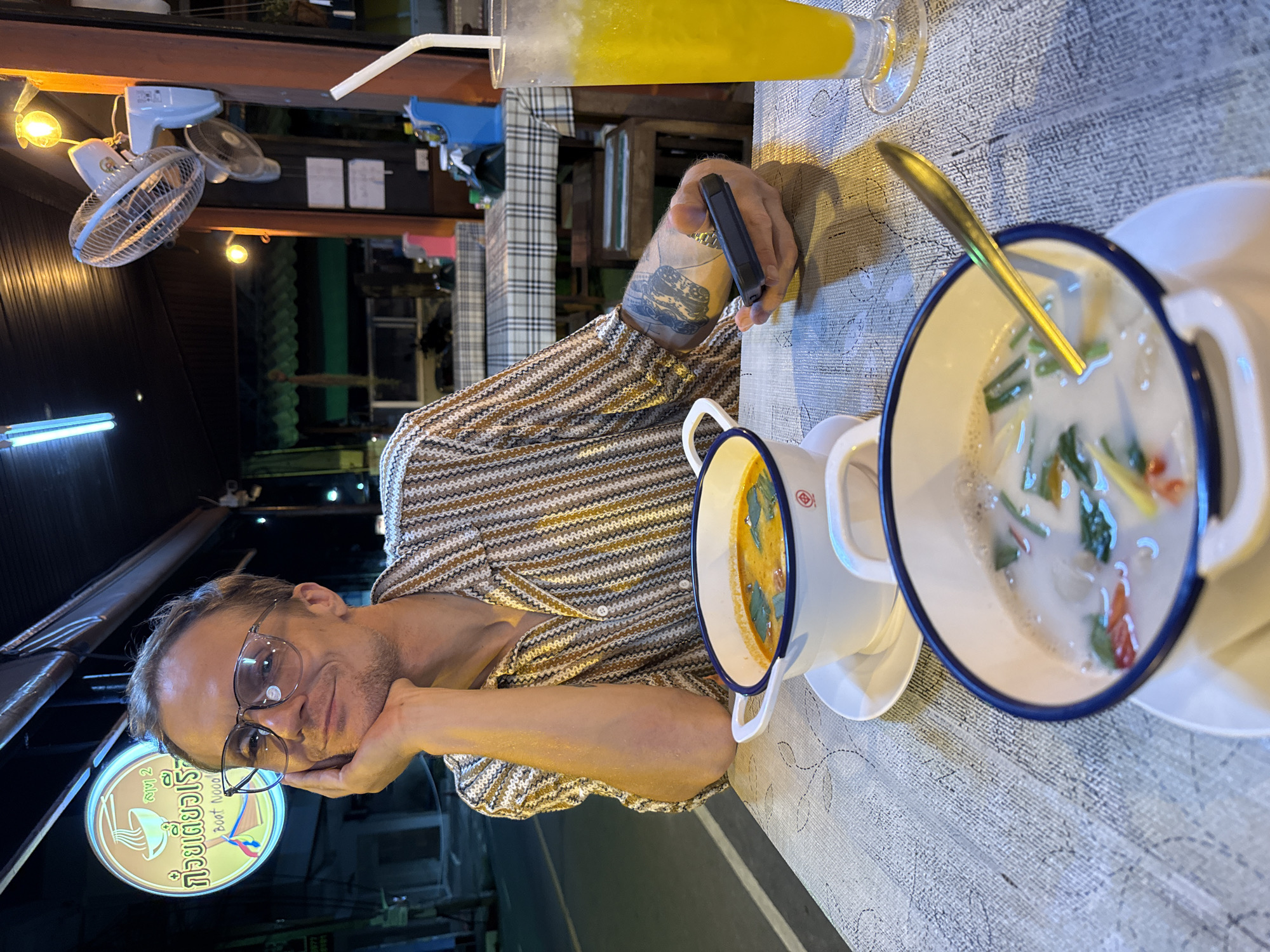 In an outdoor night setting, a man with glasses rests his head on his hand, looking at the camera. He wears a striped short-sleeved shirt and has a tattoo on his arm. In front of him are two bowls of soup, one reddish-orange and the other creamy white with green garnishes, and a glass of yellow liquid with ice and a straw. The scene appears to be at a street-side restaurant, suggested by the neon sign with Thai writing and a depiction of noodles, and a fan mounted on a post in the background. The tables are covered with checkered cloths.
