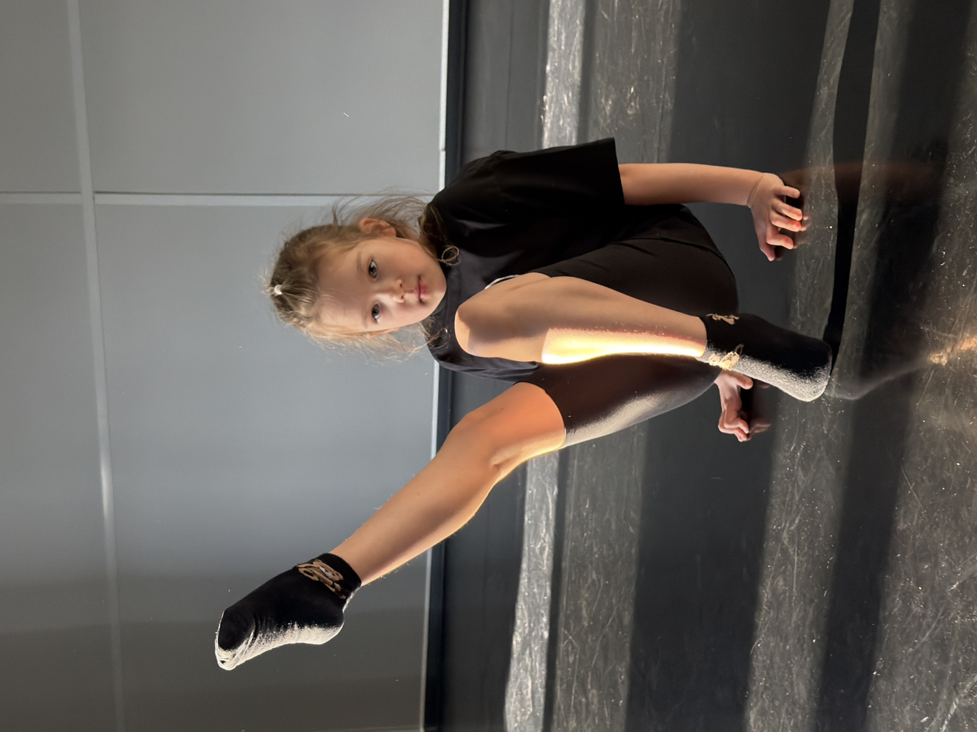 A young girl wearing black attire, including a t-shirt, leggings, and socks, is positioned on what appears to be a dark, glossy floor. Her left leg is extended towards the left side of the frame, while her right leg is bent. Her hands are placed on the floor near her body. The background consists of a light grey wall with vertical lines. The floor reflects the light and the girl's form. The photograph was taken at an angle, giving a unique perspective of the subject. There is no indication of this image being in Saint Petersburg, Russia.