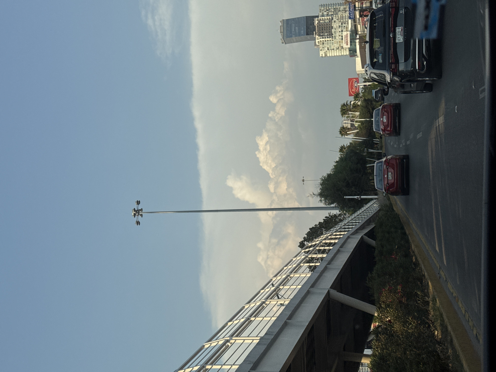 The image shows a street scene in Puebla, Mexico, with multiple lanes of traffic. Two red cars and a black SUV are visible in the right-hand lanes. A Coca-Cola billboard and several buildings are visible in the background. The foreground is dominated by a pedestrian overpass with a covered walkway. A tall light pole is situated beside the overpass. The sky is light blue, with some clouds.