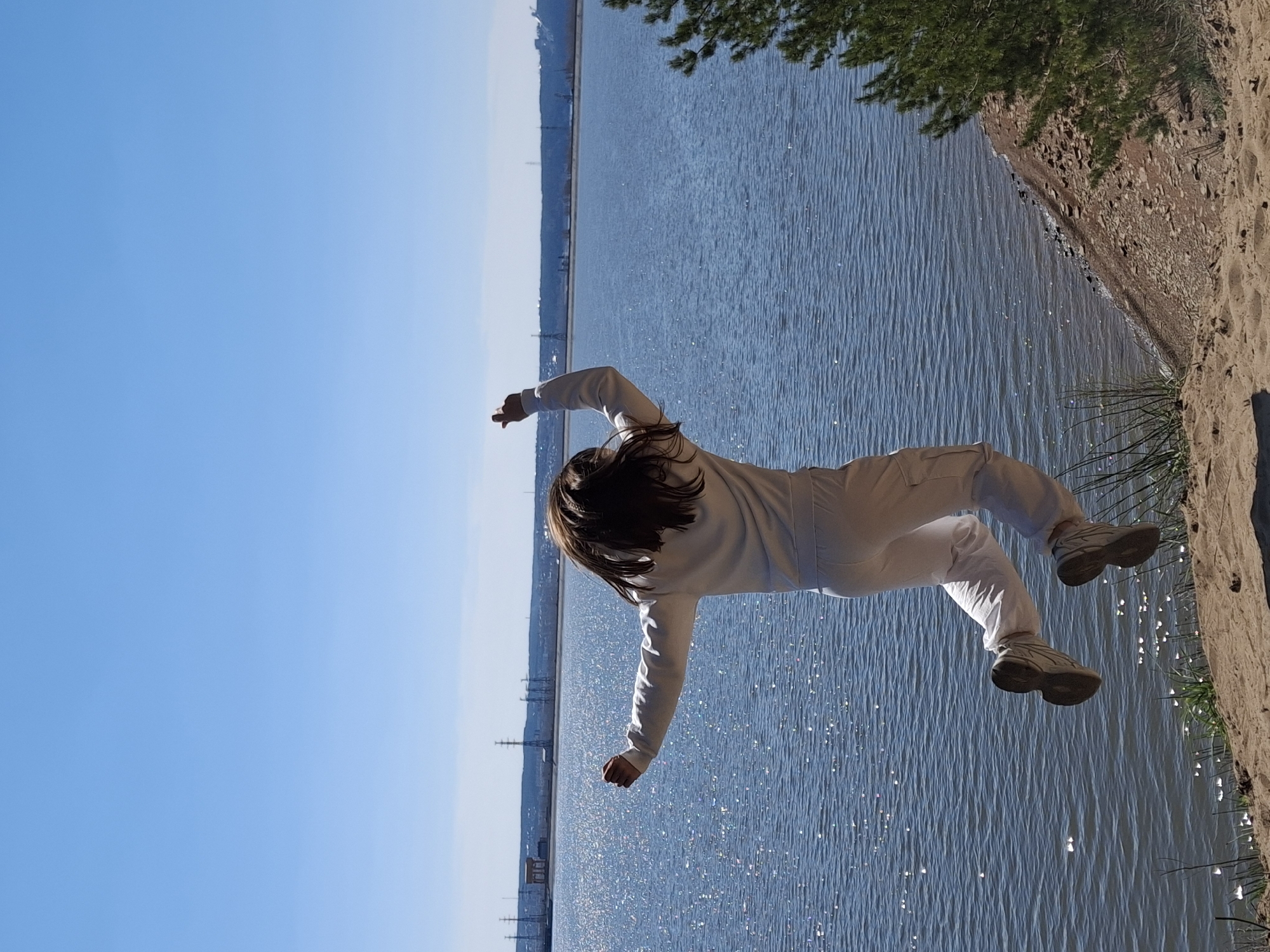 A person in white clothing is suspended in mid-air against a backdrop of water and sky. The water reflects sunlight, creating a shimmering effect. The shoreline is visible, with trees and foliage. In the distance, there are structures resembling industrial buildings or towers, potentially in Noviy, Russia. The person's posture suggests either jumping or being in a falling motion.