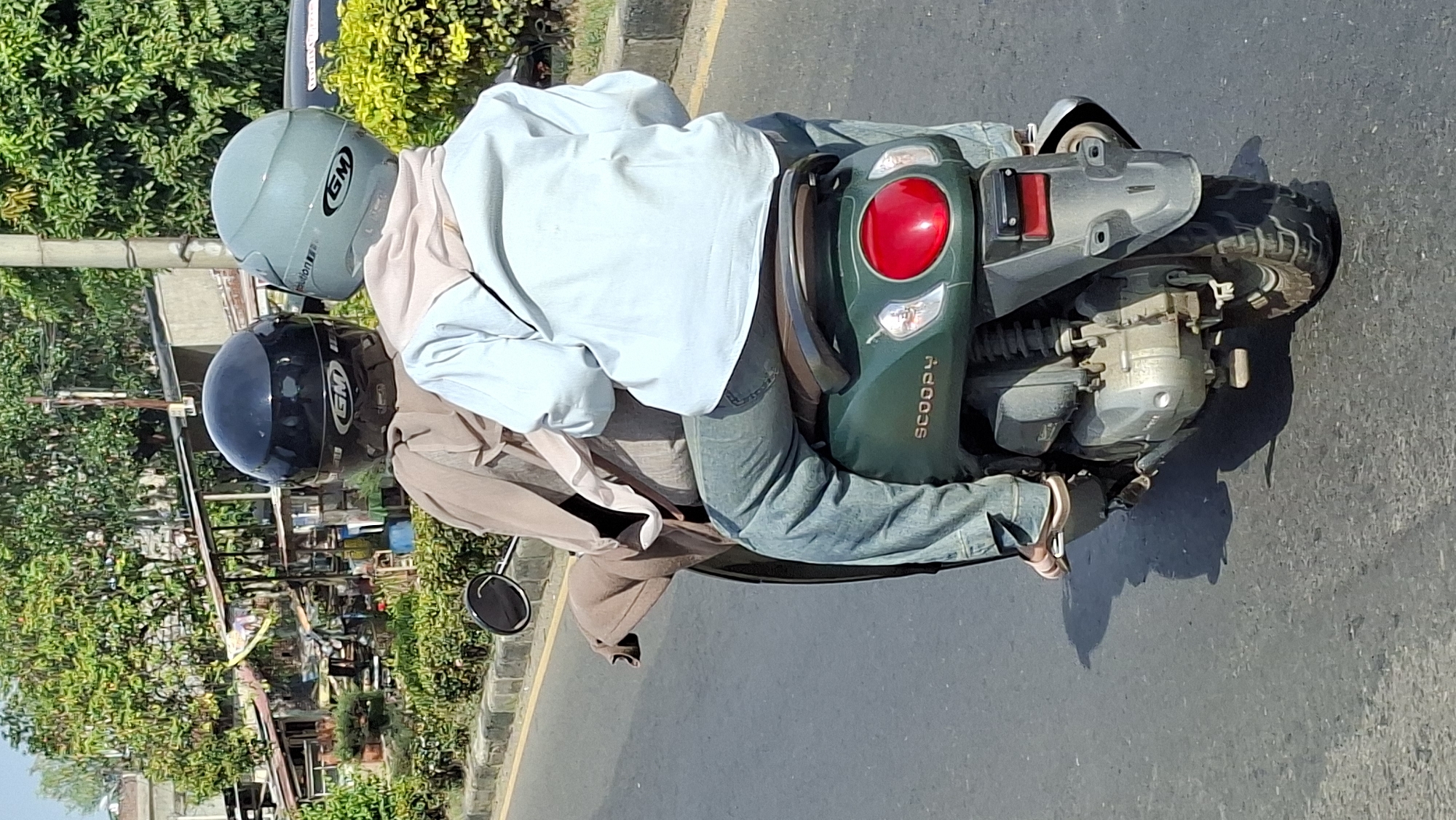 Two individuals, one driving and one passenger, are riding a dark green Honda Scoopy motorcycle on a paved road. The driver wears a black helmet with a white "GM" logo and a tan cloak. The passenger wears a light blue helmet with a white "GM" logo, a light pink scarf, a light blue shirt, and blue jeans. Both are looking forward. The motorcycle has a prominent red taillight and the word "SCOOPY" visible on its rear. The background shows a street lined with trees and small buildings, indicative of a city environment. The scene is likely in Lhokseumawe, Indonesia, based on the visual context of the street and architecture.