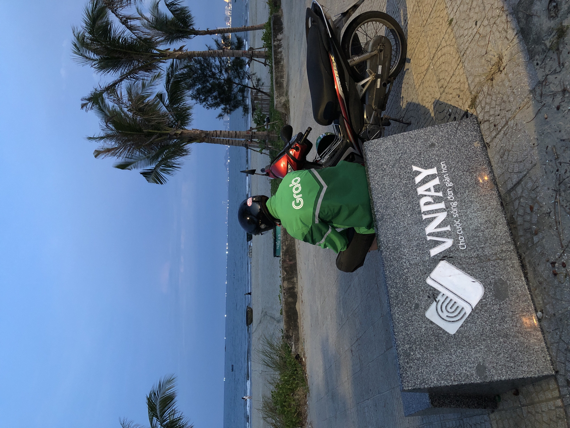 A person wearing a green Grab jacket and a black helmet sits on a stone bench facing away from the camera. A red and black motorcycle is parked to their right. In front of the bench is a large stone structure with the VNPay logo and Vietnamese text. The background shows a beach, the ocean, and palm trees against a twilight sky. The scene is located on the beach in Đà Nẵng, Vietnam.