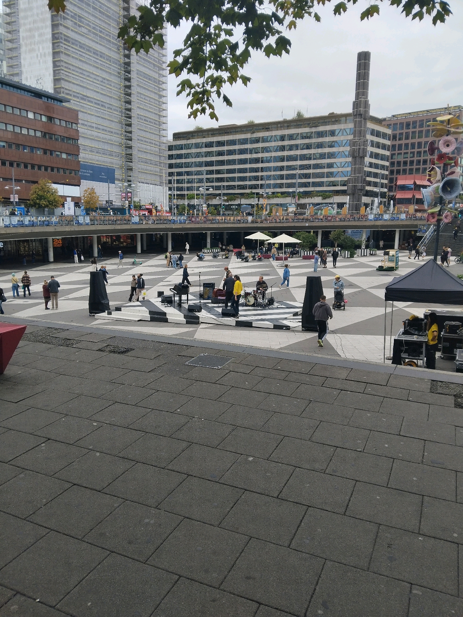 The image depicts a public square in Stockholm, Sweden, likely Sergels Torg, characterized by its distinctive black and white patterned paving stones. In the foreground, a paved area with large, dark gray tiles shows some wear and scattered debris.
The midground features the main plaza, where a diverse group of people are present. Several individuals are gathered around a small performance area set up with a keyboard, a drum set, and audio equipment. One person appears to be playing the keyboard, while another is seated behind the drums. Microphones and speakers are visible, suggesting an ongoing musical performance or setup. A large, black, opaque screen or structure stands near the performance area.
Further into the plaza, other people are walking, standing, or pushing strollers. Two large white umbrellas provide shade for some of the attendees. A portable green food cart or vendor is also present. In the background, a tall, slender, dark gray, multi-faceted sculpture rises from the plaza.
The background is dominated by modern city architecture. To the left, a tall skyscraper with scaffolding and protective sheeting is under construction or renovation. To the right and center, several multi-story buildings with large windows and a pedestrian overpass with shops and signage are visible. The sky is overcast, suggesting a cloudy day. The overall scene captures a moment of urban life with a public event or gathering in progress.