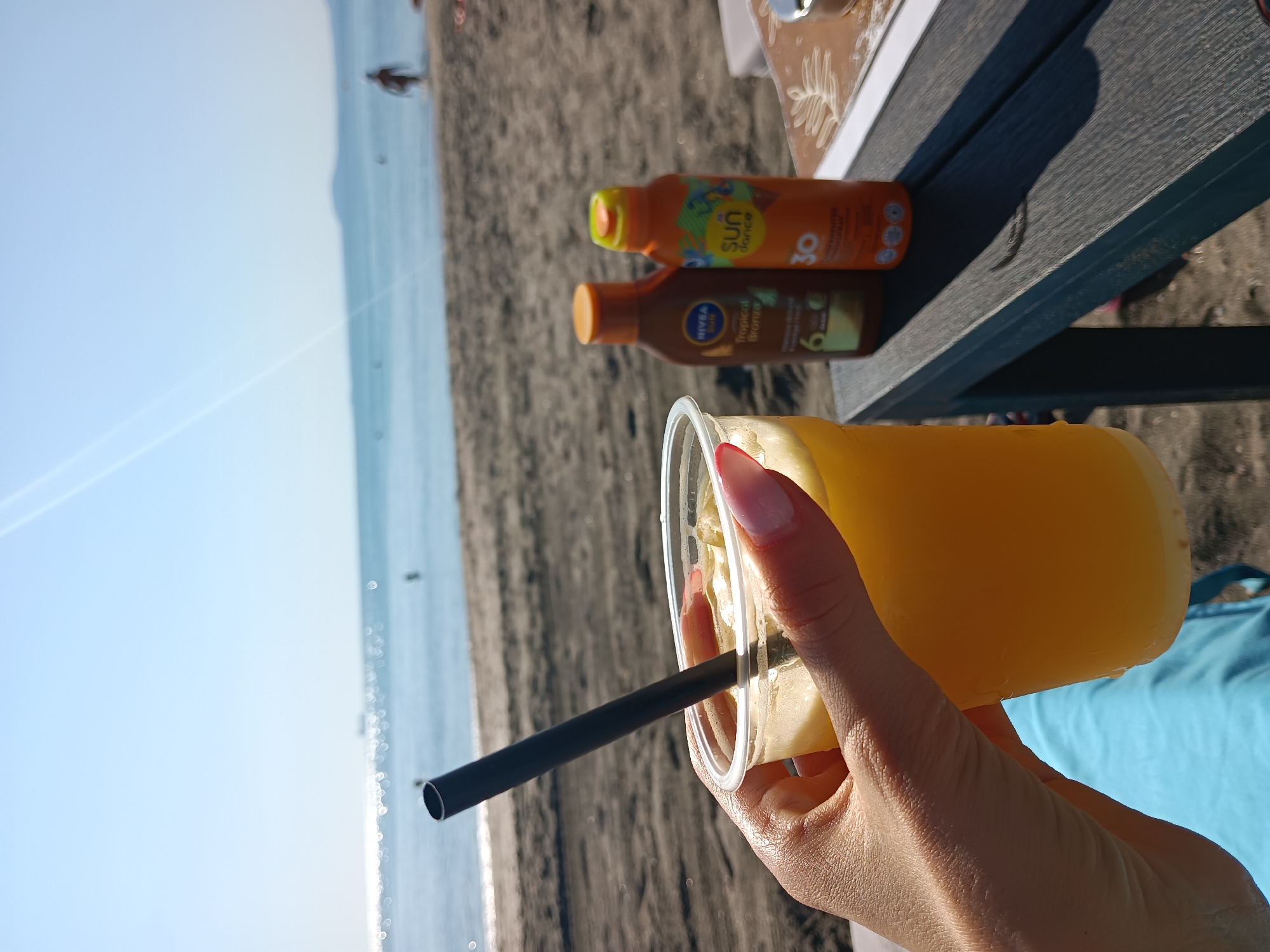 The image depicts a close-up of a hand holding a clear plastic cup filled with an orange beverage and a black straw, situated on a dark blue table. In the background, a beach scene unfolds with dark sand leading to the sea under a clear blue sky. Two sunscreen bottles, one brown labeled "NIVEA Tropical Bronze" and an orange one labeled "SUN dance 30", are on the table. The sea is visible with a distant ship and a few figures in the water. The scene appears to be a beach in Burgas, Bulgaria, given the context provided.