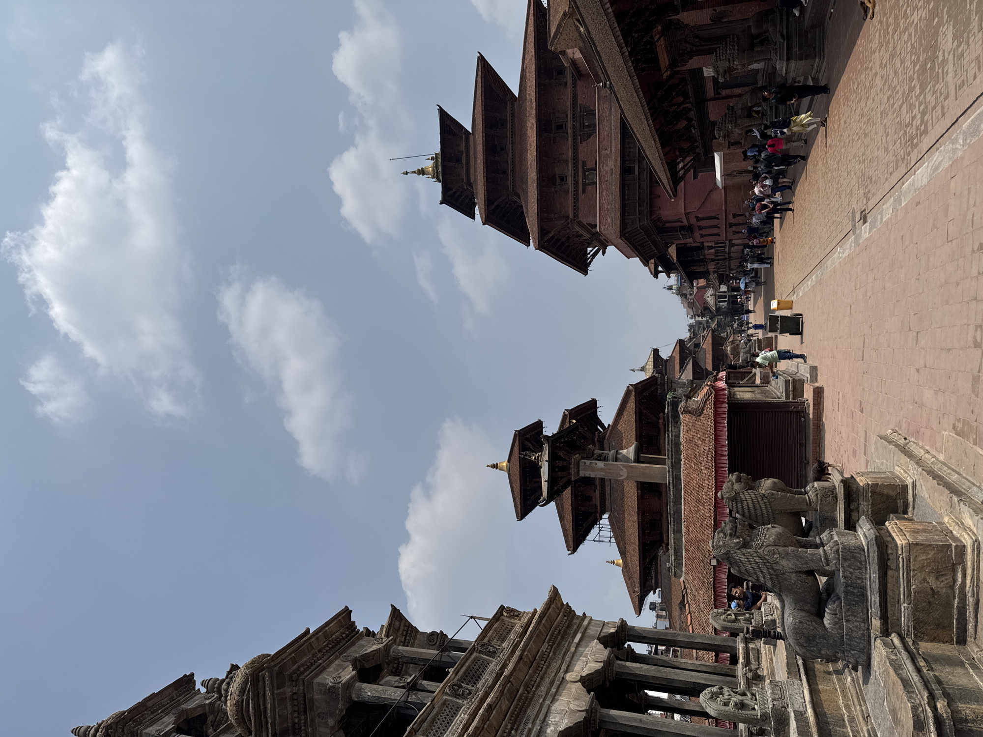 The image shows a street scene in Lalitpur, Nepal. The view is angled upward and tilted, showcasing traditional Newari architecture. Several multi-tiered wooden temples with red roofs dominate the scene, including two prominent pagoda-style structures. Intricate carvings and sculptures are visible on the temples and stone structures. On the right, a stone pathway lined with people walking and standing. The sky is pale blue with scattered white clouds. A pair of stone lion statues guard a temple entrance.