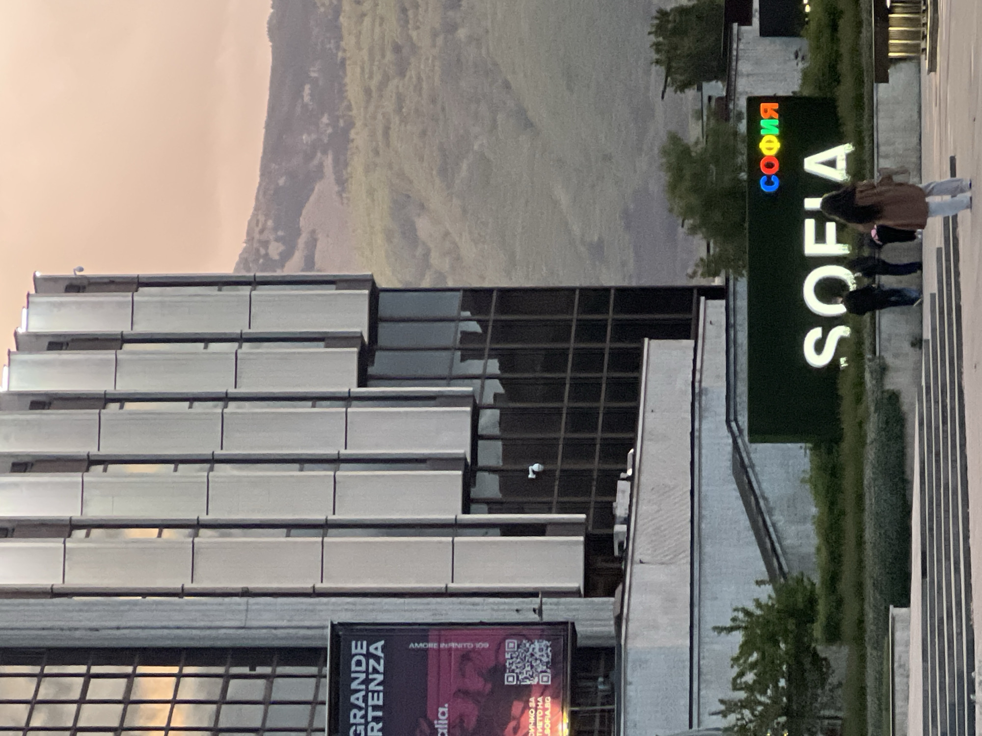 The image depicts a daytime scene in Sofia, Bulgaria, with a view of the National Palace of Culture and the Vitosha Mountain in the background. In the foreground, a multi-story building featuring a facade of horizontal rectangular panels and glass windows is visible. An advertisement with text written in Italian is attached to the building, along with a QR code. To the right of the building, a vertical sign displays the word "SOFIA" in large, white block letters. Above the word "SOFIA", the Cyrillic spelling "СОФИЯ" is rendered in multicolor (red, orange, yellow, green, cyan, blue, indigo, violet). Two people are standing on a walkway with vertical metal bars next to the sign. Vitosha Mountain rises in the background, providing a green, mountainous backdrop to the cityscape.