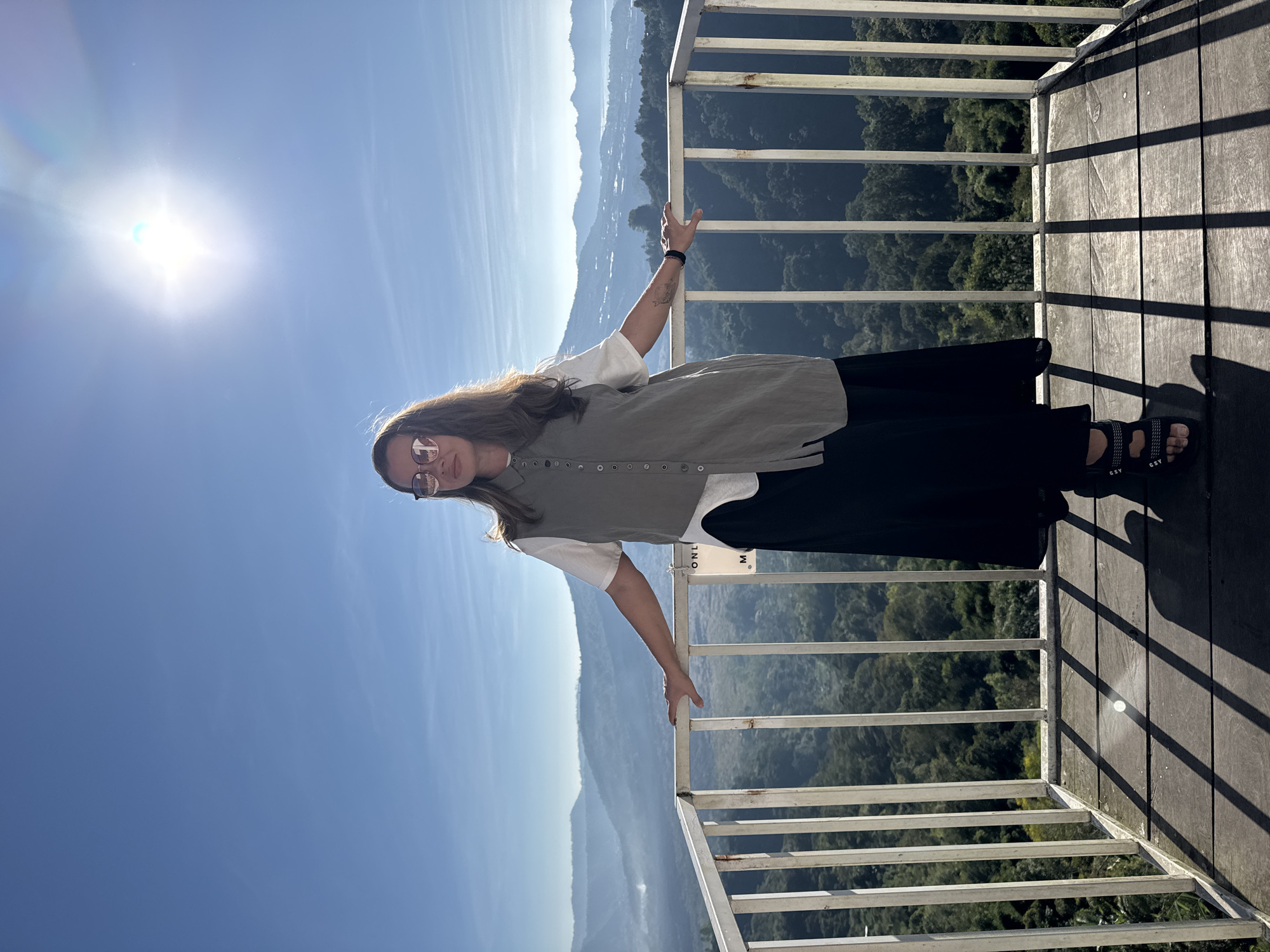 A solo adult female stands on a wooden observation deck with a white metal railing, overlooking a mountainous landscape in Kintamani, Indonesia. She is positioned centrally, facing slightly right with her head turned towards the camera, and her hands resting on the top rail of the railing. She wears sunglasses, a white short-sleeved t-shirt layered under a grey sleeveless button-down tunic, a long black skirt, and black sandals. A tattoo is visible on her right inner forearm, and a black bracelet is on her right wrist. A white sign reading "ONLY M" is partially visible on the railing to her left. The deck floor is made of wooden planks, with strong diagonal shadows cast by the railing bars. In the background, multiple layers of verdant, forested mountains and hills recede into a hazy distance. A large body of water is visible beyond the mountains on the far right. The sky is clear blue, with a bright, intense sun positioned in the upper right, causing lens flare.