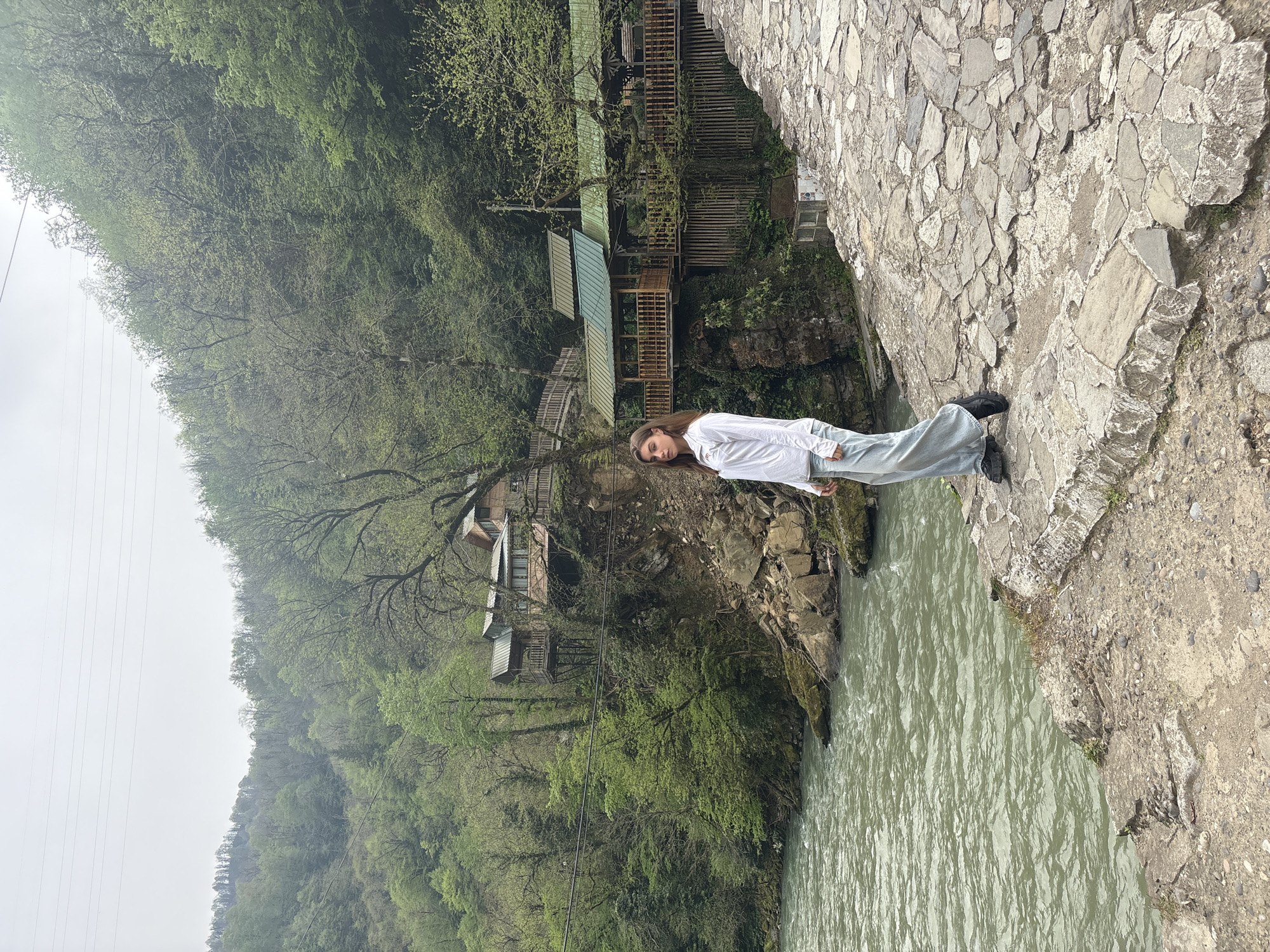 A young woman stands on a stone bridge overlooking a river in Kveda Makhuntseti, Georgia. She is dressed in a white shirt and light-colored pants, with dark shoes. The bridge is constructed of irregular, flat stones. Below, a river flows, and on the opposite bank, a lush, green hillside rises. Wooden structures, possibly homes or shops, are nestled among the trees. The sky is overcast, giving the scene a muted, natural light.