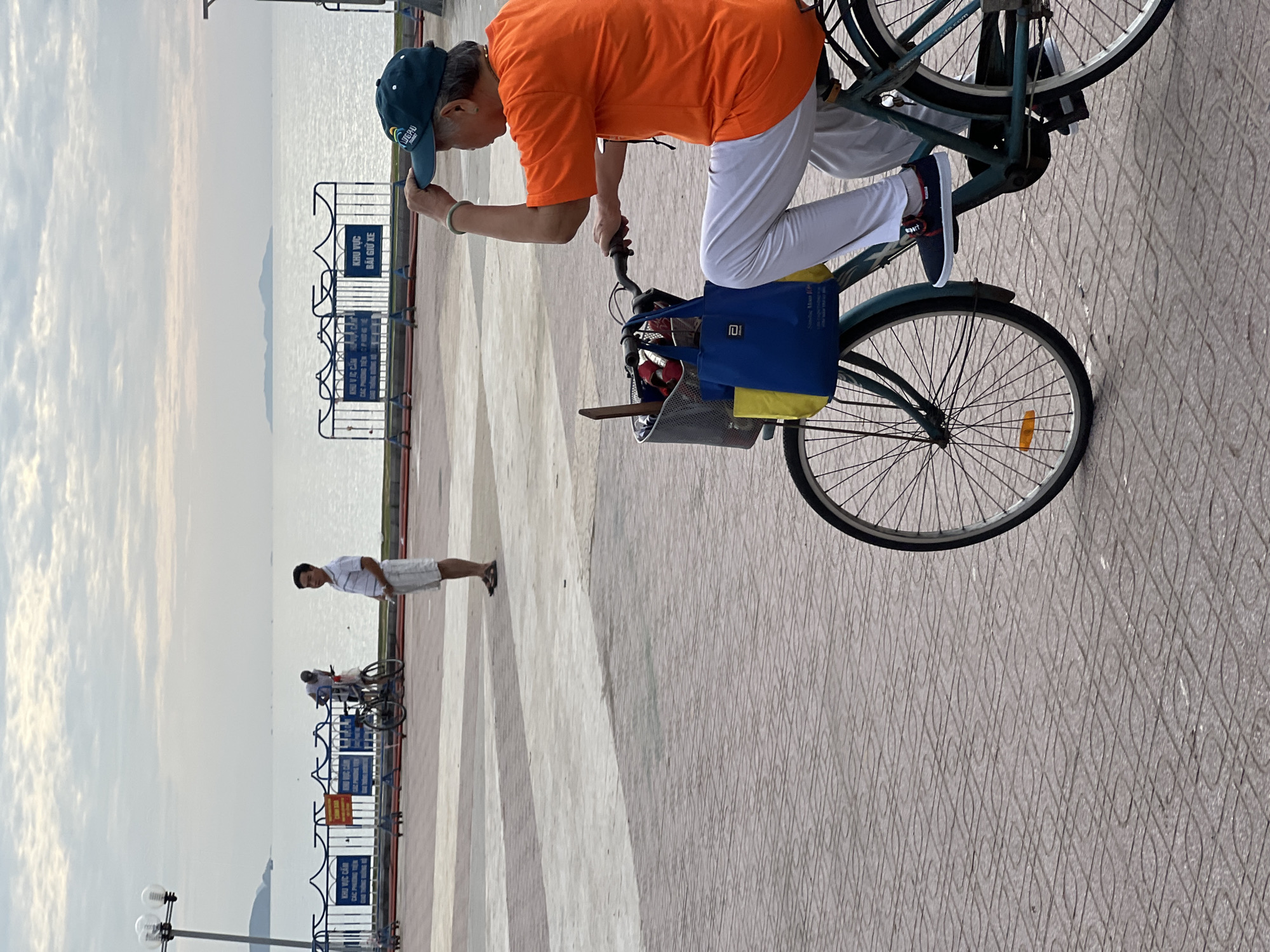 In Nha Trang, Vietnam, on a paved walkway, a person wearing an orange shirt, white pants, and a dark blue cap is riding a bicycle. The bicycle has a basket at the front filled with items, including two shopping bags, one blue and one yellow. The person's right hand is raised, touching the cap. In the background, a man in a striped shirt and white shorts stands near a bicycle. Further in the distance, a body of water stretches to the horizon under a cloudy sky, with a faint island visible. A fence with signs is positioned between the walkway and the water. The walkway is made of interlocking square tiles in various shades of beige and gray.