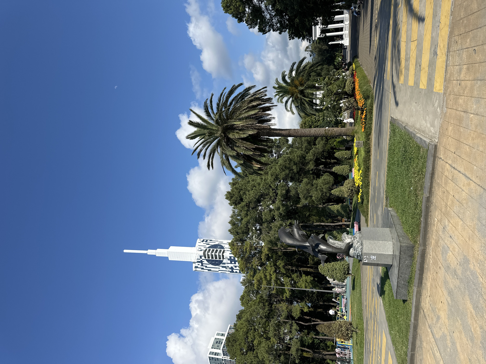 This is a photograph of a public park in Batumi, Georgia, on a clear day with a bright blue sky and scattered clouds. In the foreground, there is a statue of dolphins leaping from the water, mounted on a granite pedestal. Behind the statue, a paved pathway is visible with yellow crosswalk markings. To the right, a large palm tree stands prominently, with other palm trees and lush green trees forming a dense backdrop. Further in the background, several buildings are visible, including a tall, modern skyscraper with a distinctive white spire and a facade featuring a geometric pattern of black and white squares. To the far right, a white colonnaded structure, possibly a pavilion or part of an architectural complex, is partially visible. Scattered people can be seen in the distance, some near playground equipment on the left side of the park. The overall scene suggests a well-maintained urban park environment.