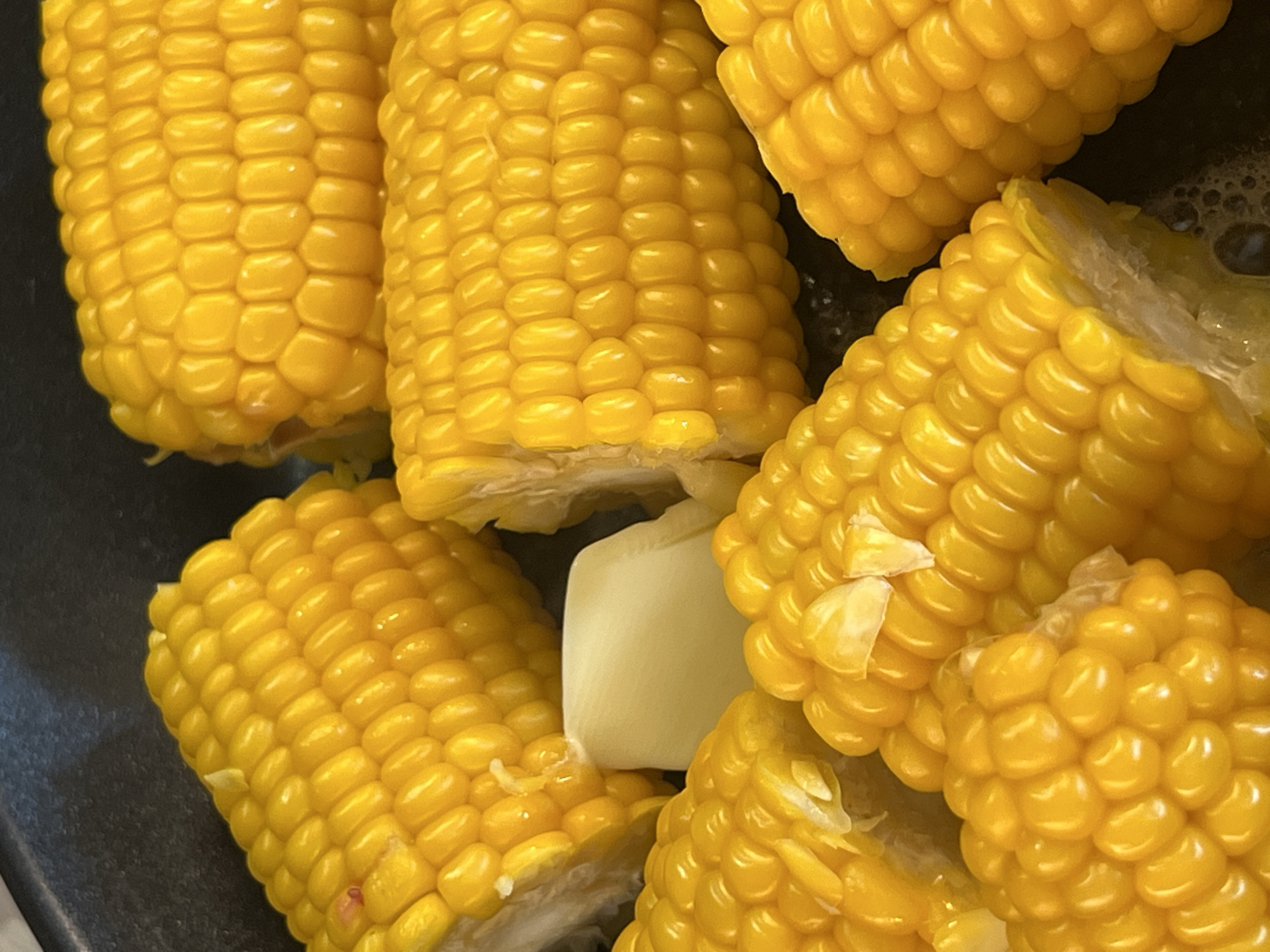 The image shows a close-up view of several pieces of corn on the cob. The corn is bright yellow and appears to be cooked, with some pieces showing glistening kernels. There is also a small piece of white butter or cheese in the center of the image. The corn is arranged in a circular pattern within what appears to be a dark pan, possibly a frying pan or pot. The background is dark and out of focus. There is no indication of the location in the city of Moosburg, Germany, within the image itself.