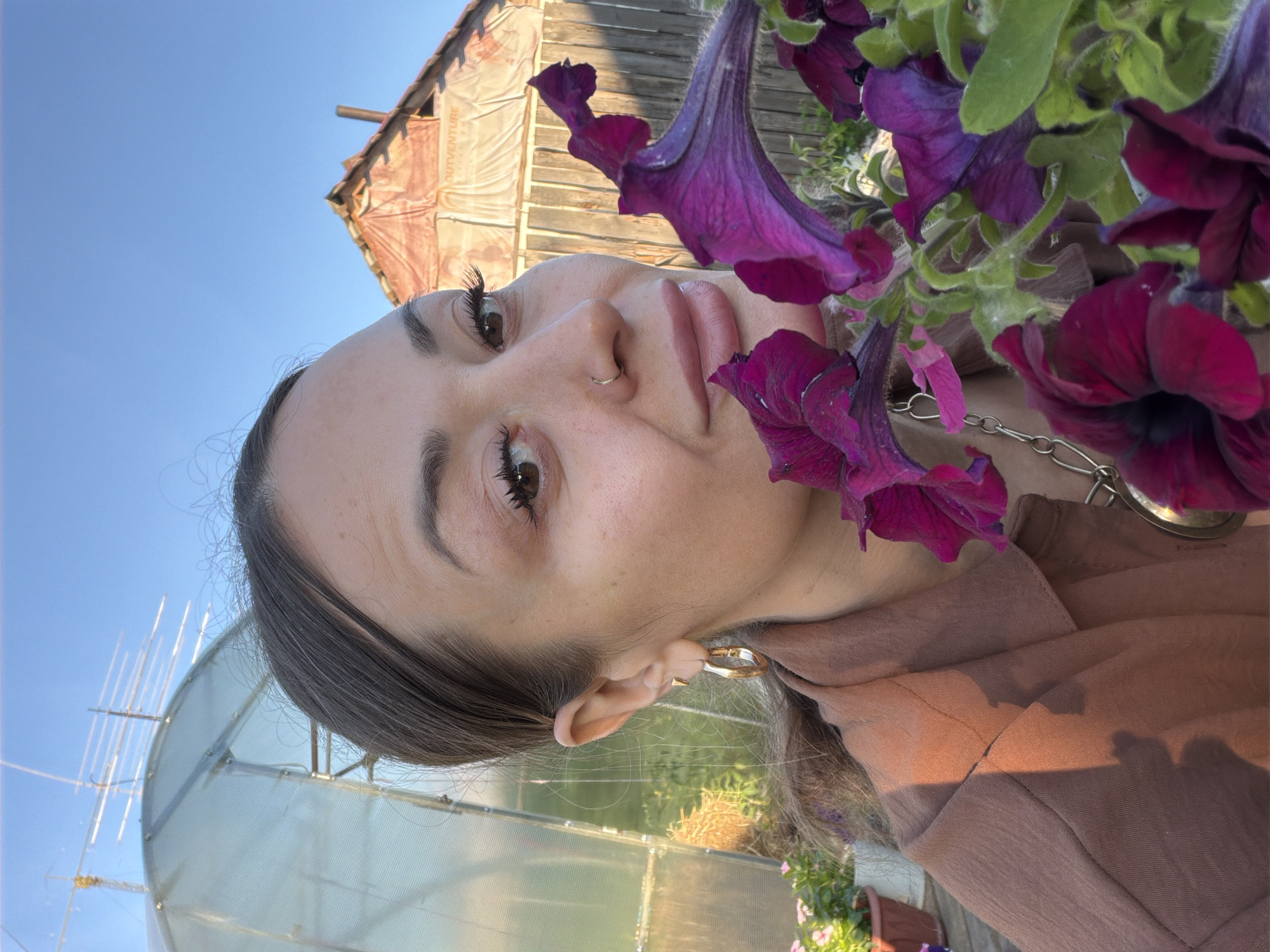 The image is an over-the-shoulder, selfie-style shot of a young woman with dark hair and brown eyes. She is wearing a brown top and a silver chain necklace. Her face is tilted upwards, with purple petunias framing her face. She has a nose ring and wears gold hoop earrings. The background shows a rustic wooden building with a corrugated roof, a greenhouse, and a clear blue sky, suggesting an outdoor setting. The lighting indicates it is daytime, likely late afternoon or early evening, with the warm glow of the sun casting shadows on her clothing and the building. The overall mood is relaxed and natural. The image does not contain any visible text.