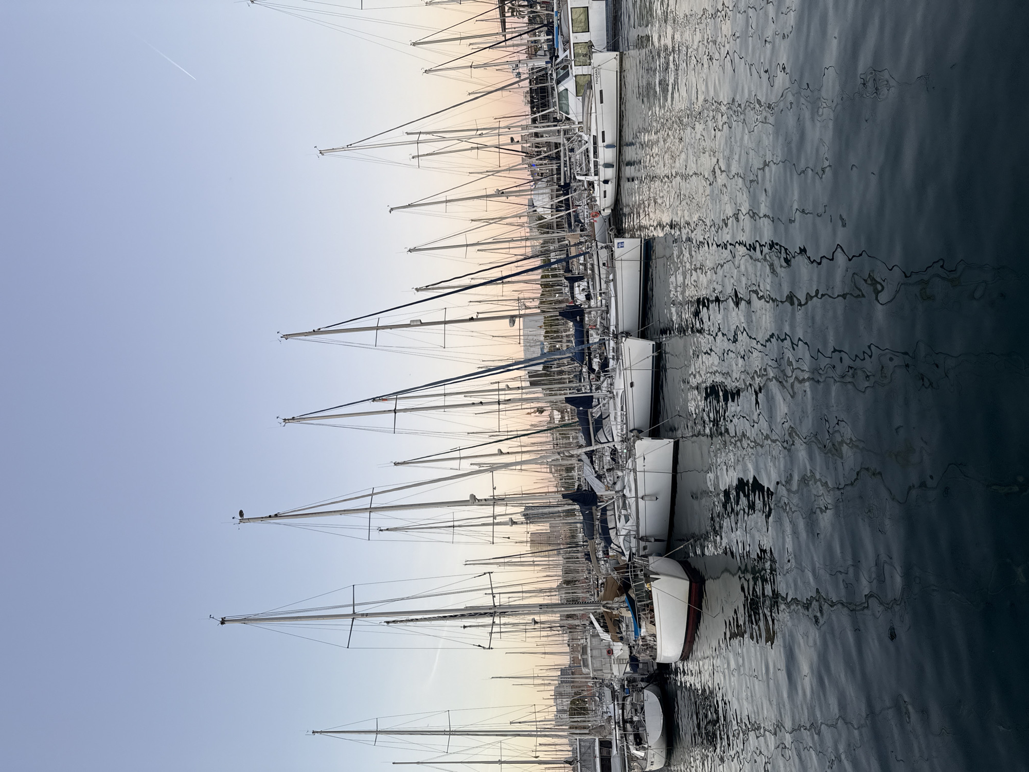 The image captures a serene view of a marina filled with sailboats at dusk in Barcelona, Spain. The sky is a soft gradient of pale blue and pink, with a faint contrail visible in the upper right corner, indicating the presence of air traffic. Numerous sailboats, mostly white with tall masts, are moored closely together in calm, dark water that reflects the boats and the sky. The reflections are distorted by gentle ripples on the water's surface, creating abstract patterns. In the background, the silhouettes of city buildings are faintly visible against the twilight sky, suggesting the marina is located near the urban center. There are no people or specific actions depicted; the scene is a still, tranquil composition of nautical elements and the ambient light of early evening.