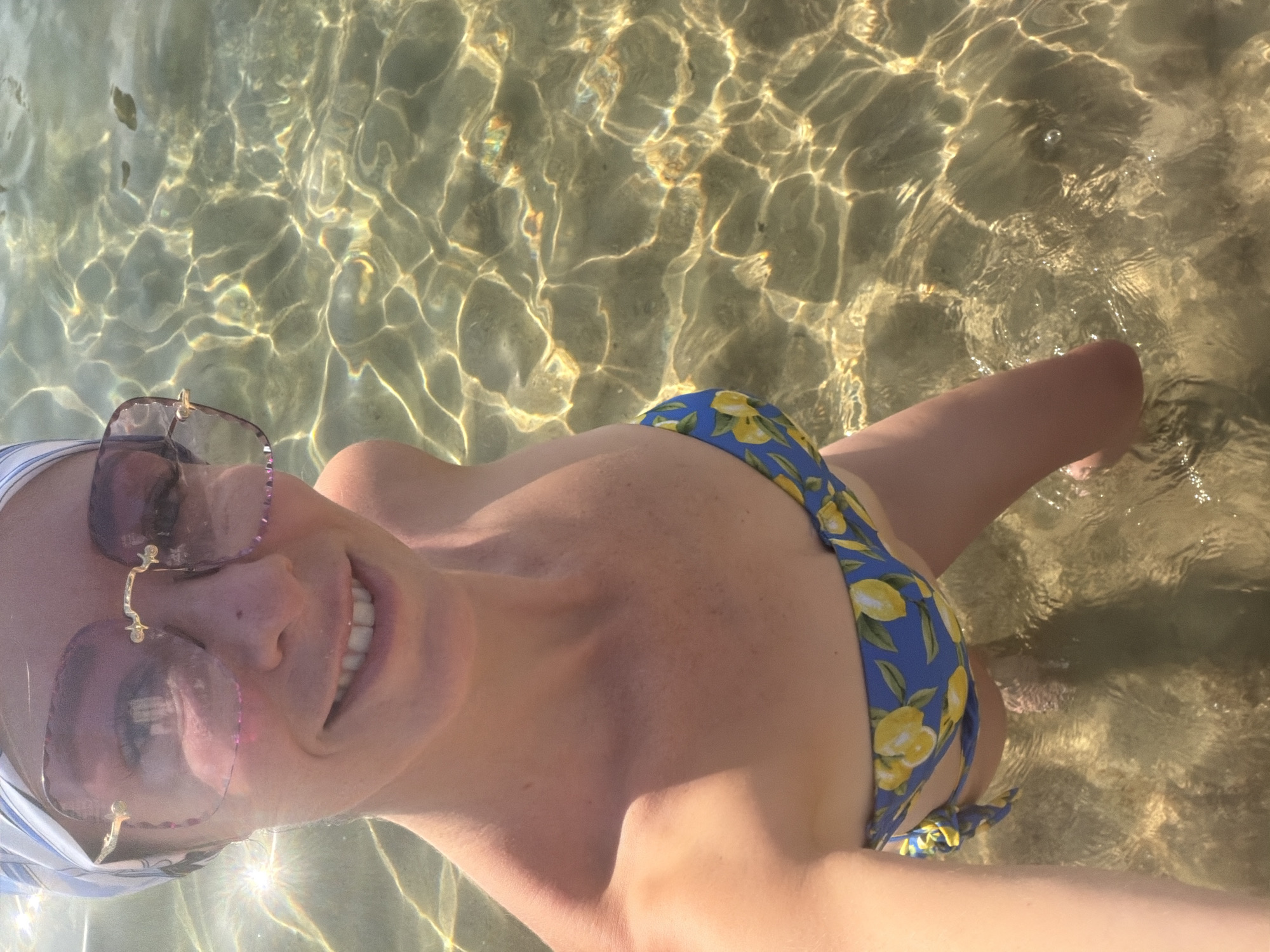 This is a medium shot, over-the-shoulder selfie of a person in shallow, clear water with a sandy bottom. The person is wearing a blue bikini with a yellow floral pattern, sunglasses with pinkish-purple lenses and gold accents, and a white and blue striped headband. The water is illuminated by sunlight, creating shimmering patterns on the sand. The person is smiling at the camera, appearing happy and relaxed. The image suggests a sunny day in a tropical location, likely Nha Trang, Vietnam, given the context. The water appears warm and inviting, and the overall mood is one of leisure and enjoyment. There is no visible text in the image.