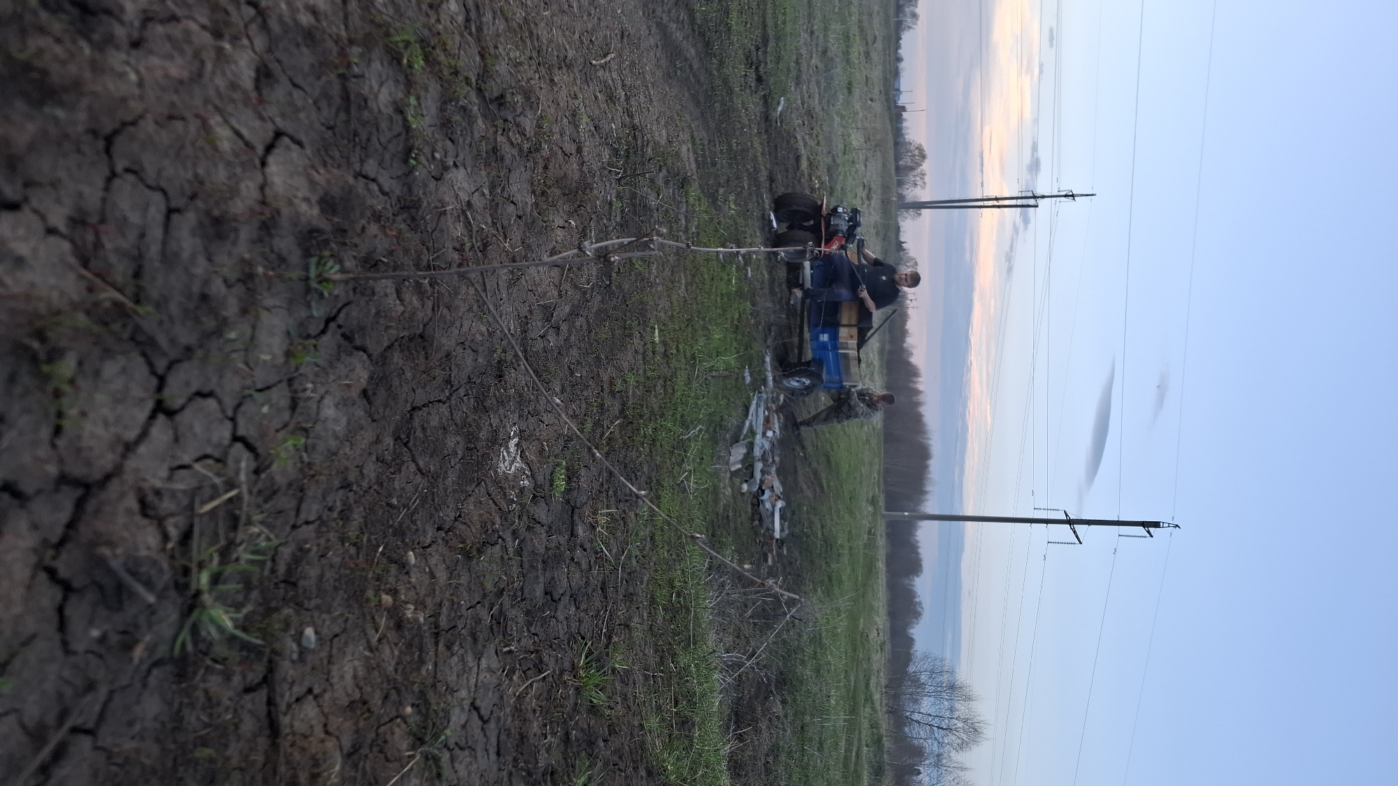 Two men are next to a small, blue four-wheeled vehicle in a field. One man is standing near the vehicle, and the other is on it. There are some pieces of material or debris on the ground near the vehicle. A young tree sits left of the vehicle, and the ground is cracked. Power lines stretch across the sky. The sky is light blue near the horizon with some pink and orange hues. The location is Dubrova, Russia.