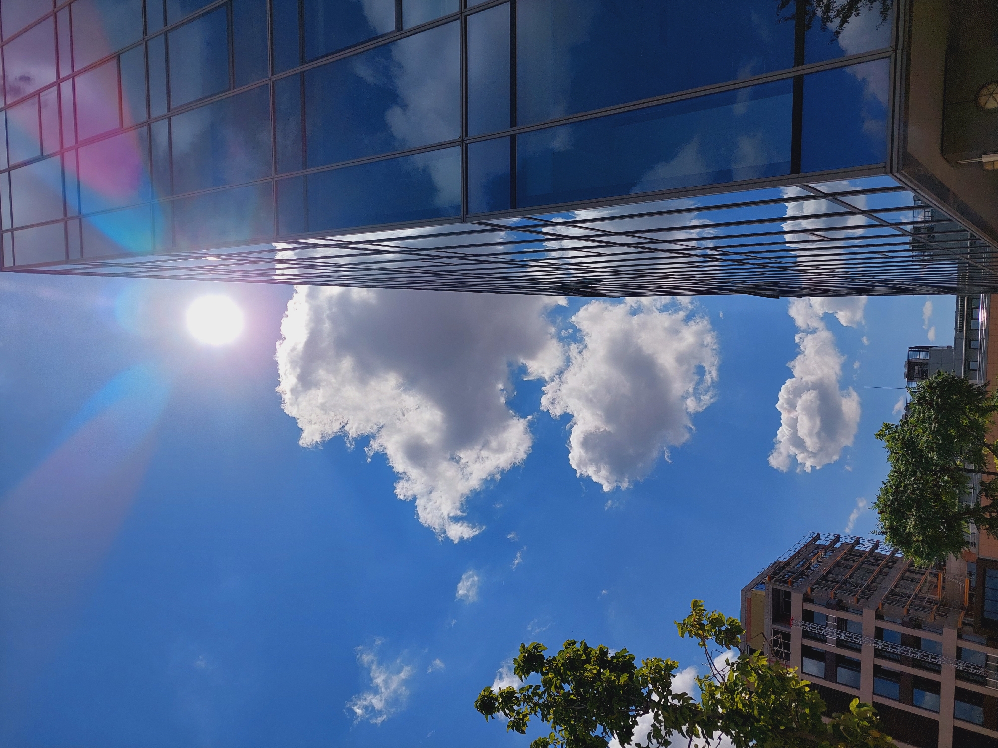 The image captures a low-angle view of a modern glass-fronted building in Sofia, Bulgaria, under a bright, sunny sky. The sun is visible, emitting a strong glare and lens flare, with light blue and pink hues radiating outwards. The building's facade is made of reflective glass panes, mirroring the blue sky and white, fluffy clouds.
To the left of the glass building, another building, possibly under construction, is visible. It has a lighter colored facade and scaffolding, with dark-windowed sections. Green trees with lush foliage are in the foreground, partially obscuring the lower parts of the buildings and providing a contrast of organic shapes against the geometric architecture. The sky is a vibrant blue, dotted with several scattered clouds. The overall scene depicts an urban environment with a focus on modern architecture and the interplay of light and reflection.