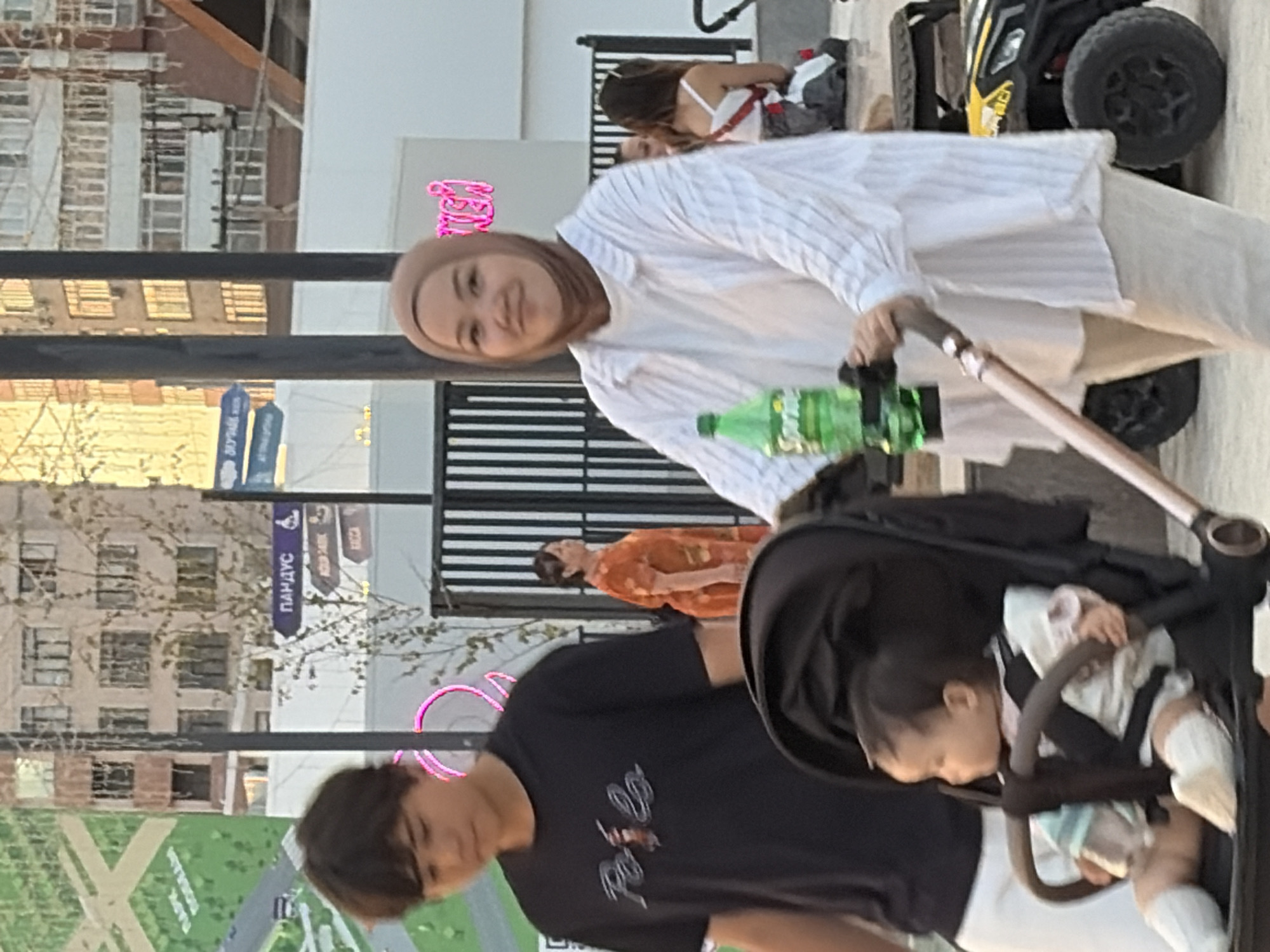 In an outdoor urban setting, a young boy walks towards the left of the frame, his head down. To his right, a woman in a light-colored hijab and a white striped shirt pushes a stroller containing a baby. The baby, wearing white socks and a light outfit, is seated and looking to the side. The woman holds the stroller handle and has a green bottle of Sprite attached to it. In the background, a woman in an orange dress stands facing away from the camera, and further back, other people are visible, as well as buildings with windows and signage. There is also a small black and yellow vehicle to the right of the frame. The scene appears to be in a public area with fences and trees.
