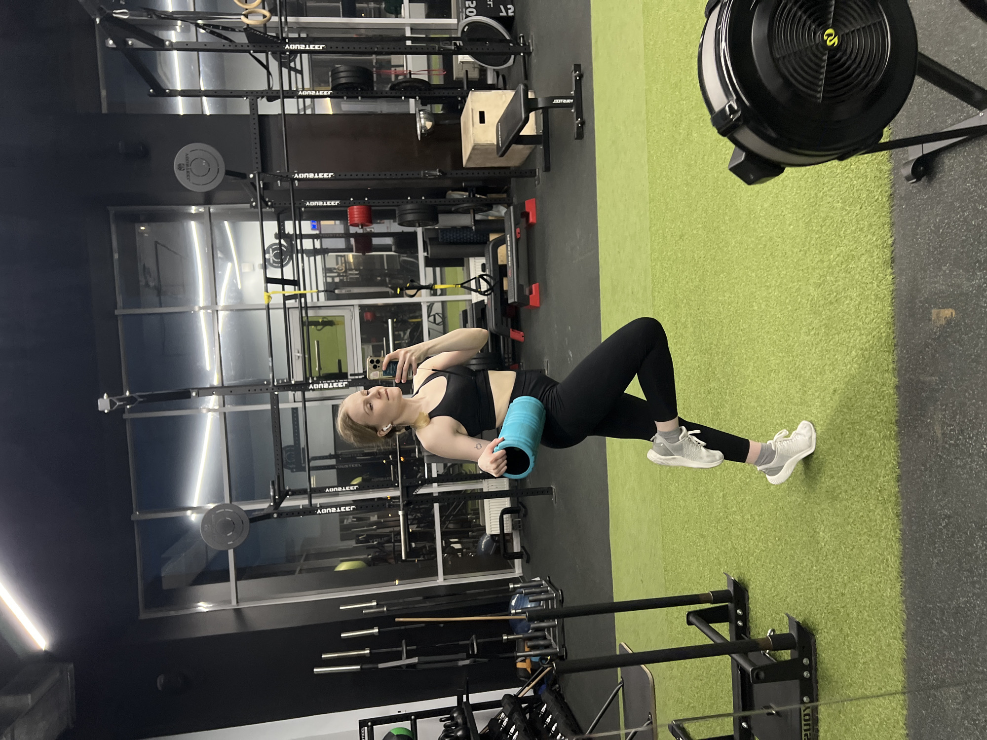 A young woman is in a gym, using a blue foam roller on her upper thighs as she poses for a selfie with her smartphone. She is wearing black leggings, a black sports bra, and white sneakers. She has fair skin and blonde hair.
The gym equipment visible includes weights, weight racks, a rowing machine, and plyometric boxes. The flooring is a combination of green artificial turf and dark rubber mats. Large windows are visible in the background.
The information about the city of Khimki, Russia, cannot be verified from the image alone.