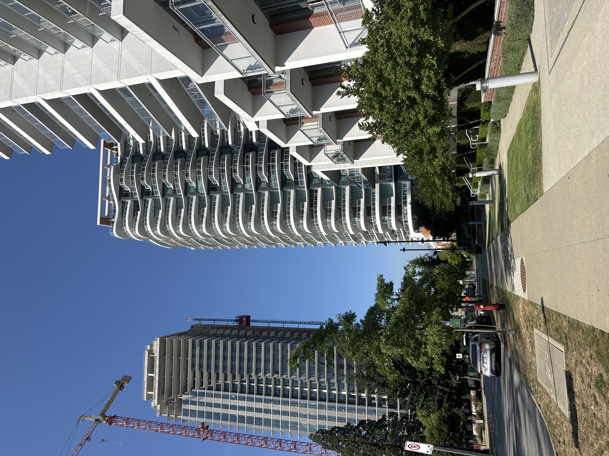The image depicts a street scene in Surrey, Canada, on a clear, sunny day. Two modern, multi-story residential buildings dominate the skyline. The building on the right is completed, with white facades and numerous balconies featuring glass railings. The building on the left is under construction, a tall, rectangular structure with visible concrete framework and some exterior panels installed. A large red construction crane is positioned next to the unfinished building.
In the foreground, a paved sidewalk runs parallel to the street. Adjacent to the sidewalk is a strip of grass with several trees providing shade. A silver sedan is parked along the curb on the left side of the street. A red fire hydrant is visible near the sidewalk. Further down the street, more trees and greenery are present, along with glimpses of other buildings and infrastructure. The overall impression is one of urban development and modern architecture in a residential area.