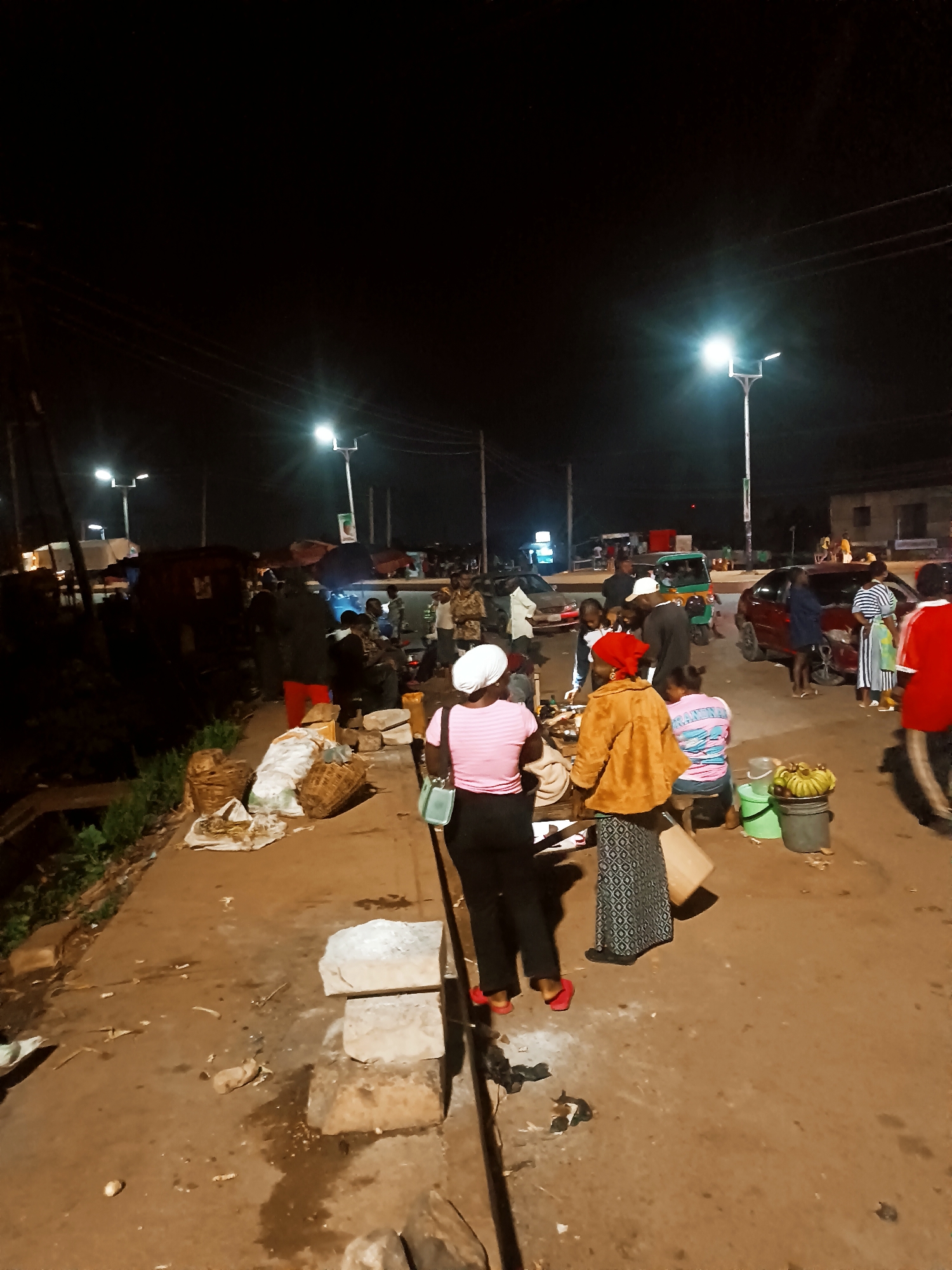 At night in Bukuru, Nigeria, an outdoor market is active under the glow of streetlights. Numerous individuals gather; some appear to be vendors selling goods like bananas displayed in buckets, while others seem to be customers. A woman in a pink shirt and black pants stands near some stone blocks, and other individuals wear various clothing. Baskets and bags sit on the ground near a drainage ditch. Several vehicles, including a red car and a small motorized tricycle, are visible further down the street. The ground is uneven and appears to be dirt and concrete mix.