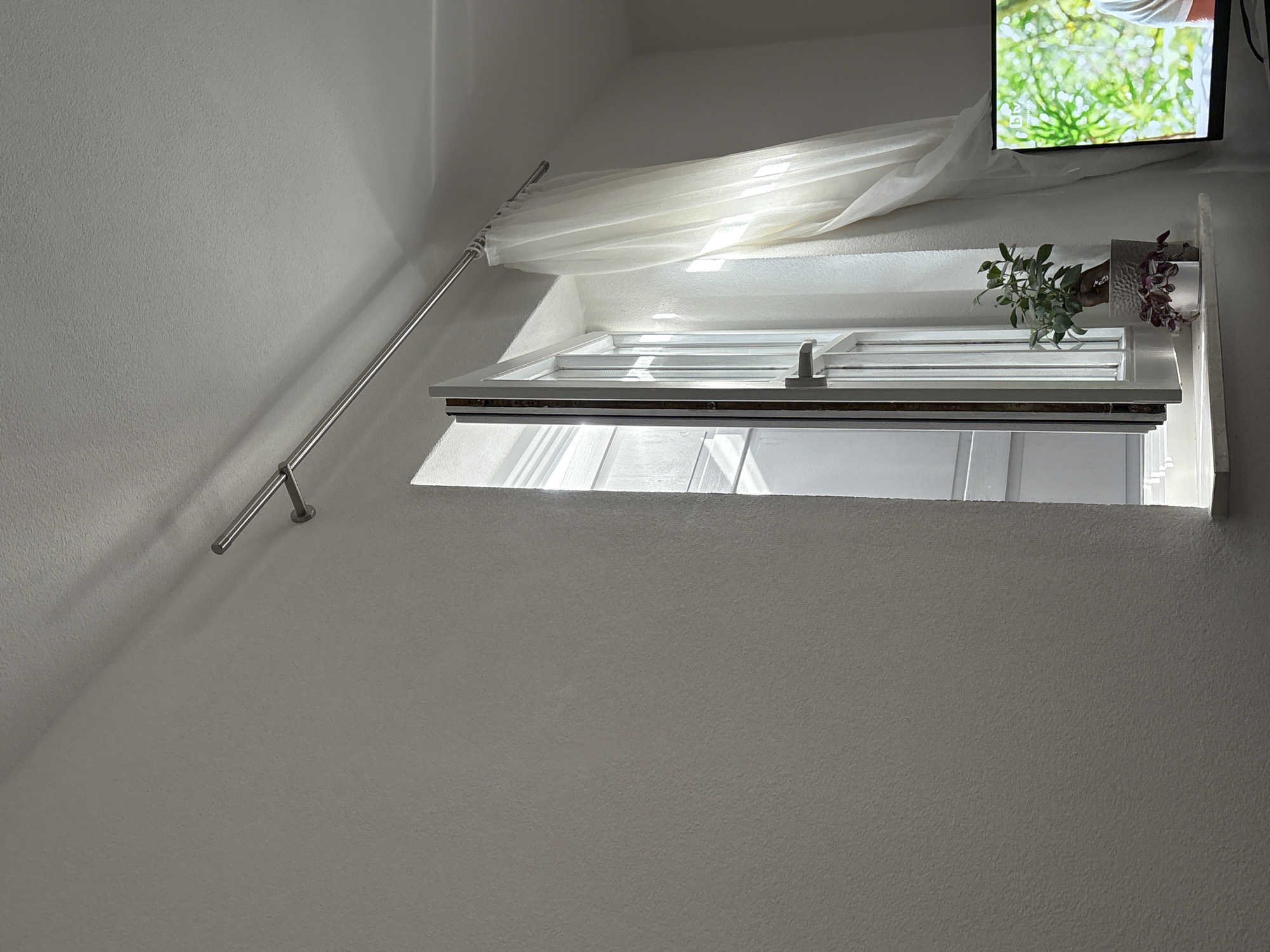 The image shows an interior view of a room with white walls and a dormer window. A metal curtain rod extends diagonally upward along the wall, supporting a sheer white curtain that partially covers the window. The window itself is white with a dark trim and is slightly open, allowing sunlight to stream in. Two potted plants, one with green foliage and another with purple leaves, sit on the window sill. A television is mounted above the window on the wall, displaying a scene with green vegetation. The room appears to be located in Forbach, Germany.