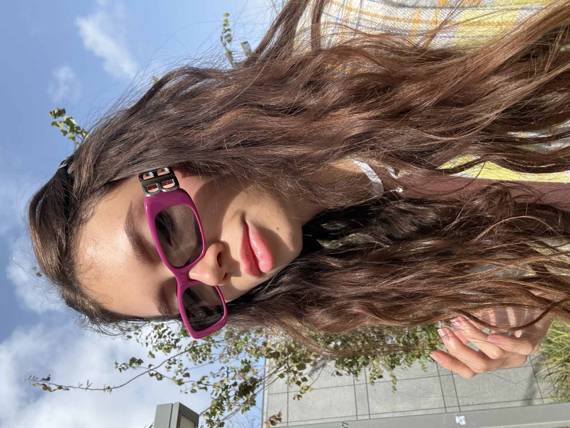 A young woman is the focal point of this medium, eye-level shot, likely taken in the city of Tel-Aviv, Israel. She is wearing oversized pink sunglasses with the Balenciaga logo on the temples, adding a touch of designer fashion. Her hair is long, wavy, and brown, contrasting with the vibrant color of the eyewear. She is wearing a brown top under a yellow plaid shirt.
The background offers visual clues to the setting. A partly cloudy sky suggests a sunny day. A tree with small, green leaves is visible. The presence of a modern building with a concrete exterior indicates an urban environment. The image suggests the woman is outdoors in a contemporary urban setting, such as Tel-Aviv, Israel.