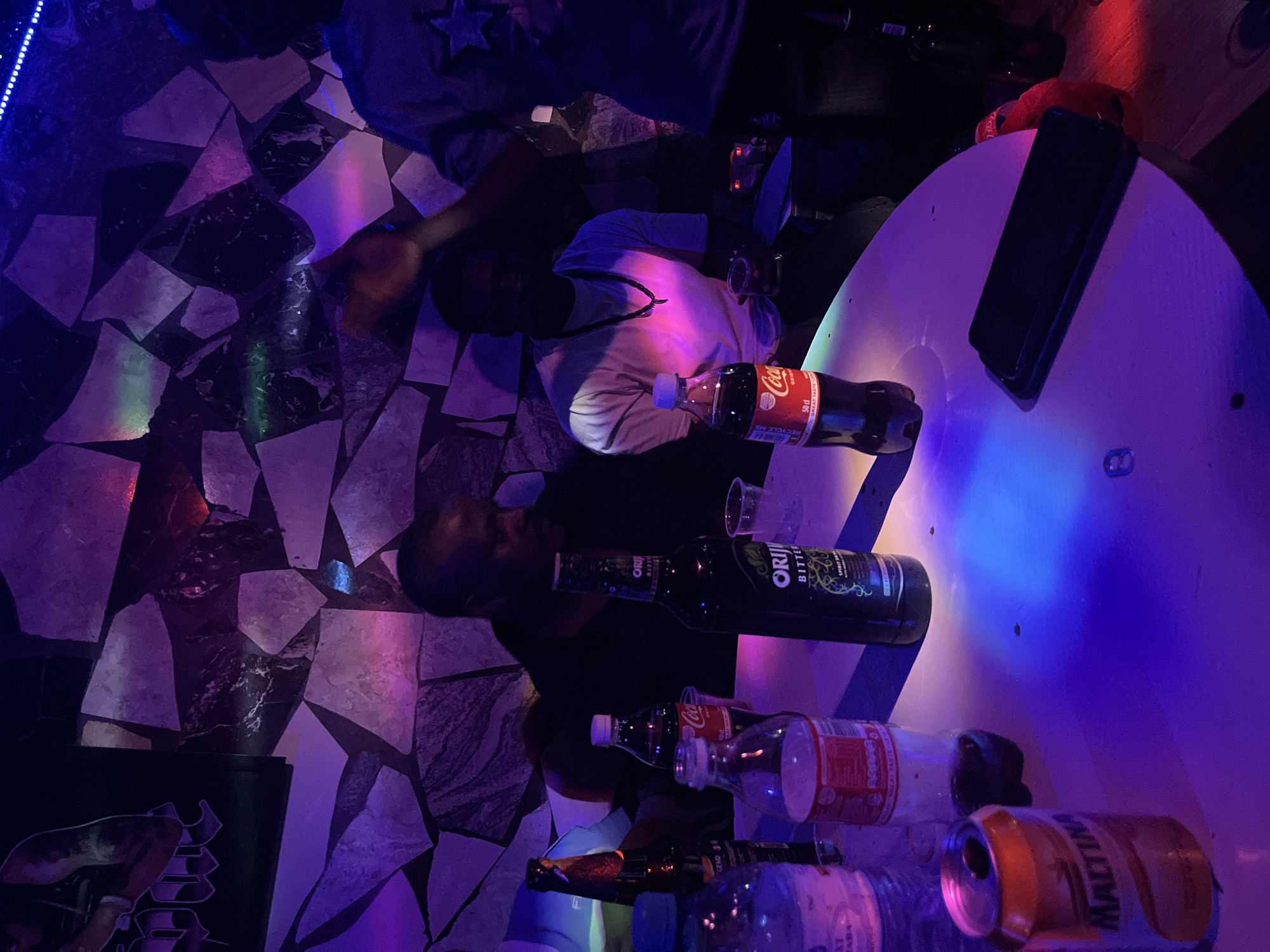 The image captures a vibrant, dimly lit indoor social gathering, likely at a bar or club, in Jalingo, Nigeria, at night. The scene is dominated by dynamic, multi-colored lighting, featuring streaks of blue, purple, pink, and green light reflecting off surfaces.
Subjects include at least two men, seen from an overhead perspective. One man, wearing a light-colored t-shirt and a dark necklace, is partially visible near a white, round table, illuminated by pink and purple light. Another man in a dark t-shirt is also seated or standing nearby. A third person's leg, clad in blue, is visible in the upper left corner.
The setting features a distinctive floor made of irregularly shaped, light and dark tiles, possibly stone or marble. A string of blue LED lights runs along the floor's edge.
On and around the white table are numerous objects, primarily beverages. These include a red bottle of "Coca-Cola" (50 cl, with "RECYCLE ME" visible), at least two dark green bottles of "ORIJIN BITTERS" (Spirit Blend), and a can of "MALTINA." Several other clear and amber-colored bottles, some empty, and clear plastic cups are also present. A black smartphone rests on the table.
The activity taking place is a casual social gathering, where individuals are enjoying drinks in a relaxed, festive atmosphere. The overall mood appears convivial and informal. No specific emotions are clearly discernible on the partially visible faces.
