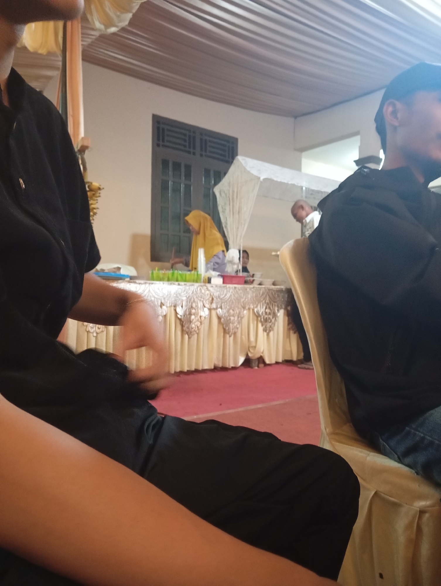The image depicts an indoor event in Kemangkon, Indonesia, likely a gathering or reception. In the foreground, a person is seated, wearing a black garment and black pants. Their left arm is visible, adorned with a bracelet. To the right, another individual is seated, wearing a black hooded jacket and blue jeans. Their head is turned away from the camera, with only their profile visible. In the background, a buffet table is set up, draped with a cream-colored cloth featuring ornate trim. On the table, various food items and drinks are displayed. A woman in a yellow hijab stands behind the table, and another person is partially visible in the distance. The ceiling is decorated with white fabric drapes. The floor is covered with a red carpet. The overall ambiance suggests a community event or celebration.