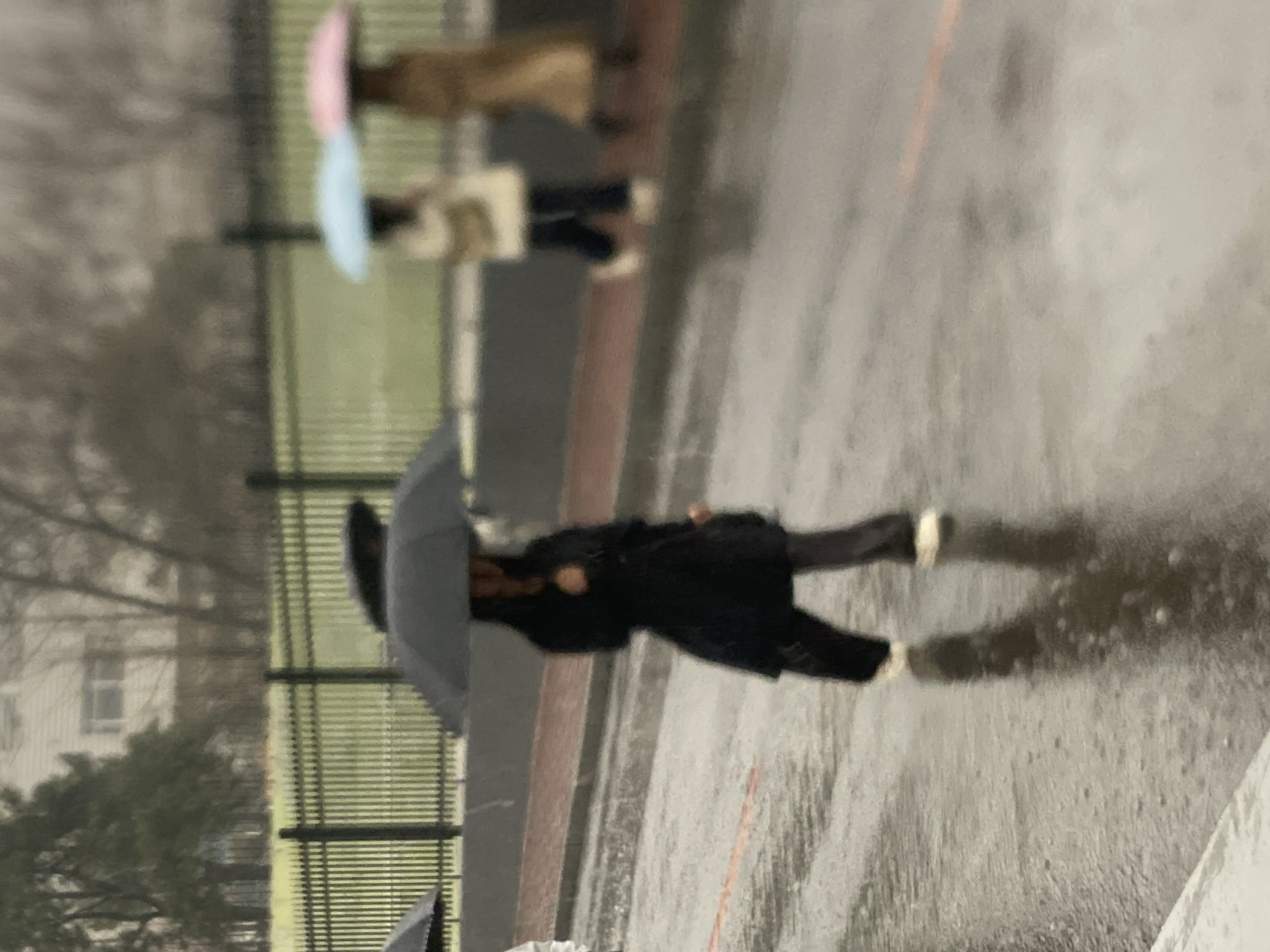 The image shows a wet, paved surface, likely a street or courtyard, with several people visible. In the foreground, an individual wearing a dark coat and dark pants, with white shoes, is walking while holding a black umbrella. The pavement is wet, showing rain droplets and reflections. In the background, there are two other people also walking with umbrellas; one has a pink and blue umbrella, and the other a neutral-colored one. A fence is visible to the left, with trees and a building further in the background. There is a street with a road-marked painted reddish orange line. Based on the information provided and the environmental conditions depicted, the location is Zhongtun, China.