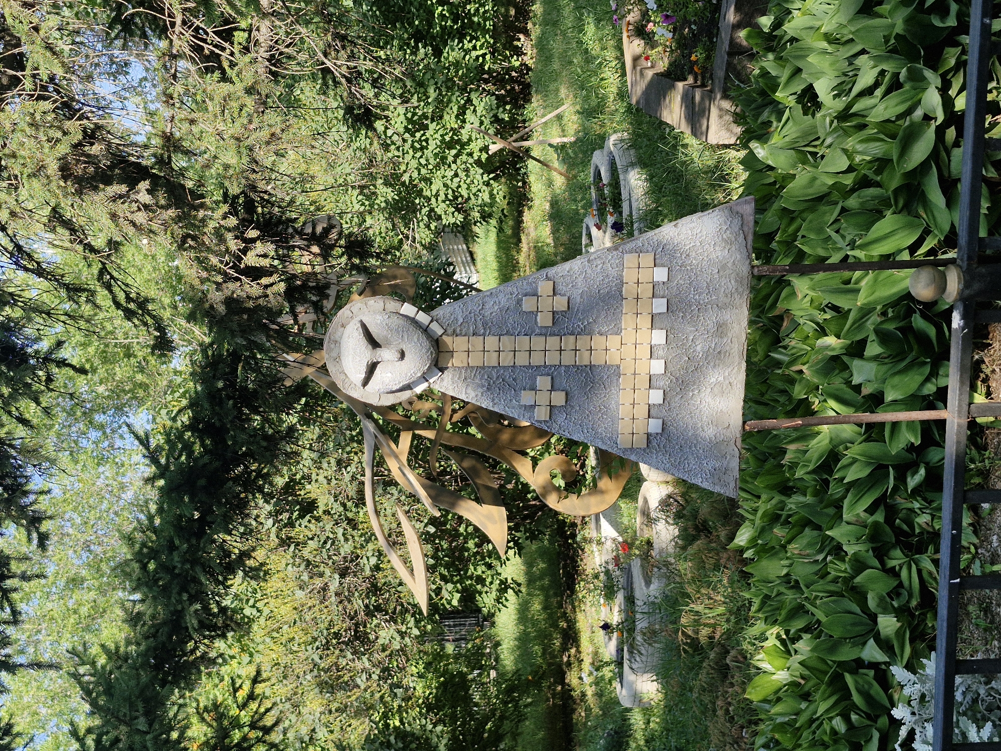 An outdoor sculpture resembling a stylized figure with a triangular body and a head with large, almond-shaped eyes stands in a garden. The sculpture's body is decorated with a pattern of small, square tiles, alternating between gold and white, forming a cross-like design. To the left of the sculpture, a large, abstract metal structure with flowing, curved elements is visible. The sculpture is supported by two thin, metal rods.
The sculpture is situated amidst lush greenery, including large, broad-leafed plants in the foreground and trees with dense foliage forming a canopy overhead. In the background, there are raised circular planters, some containing small flowers, and a wooden tripod structure. The scene is bathed in sunlight, casting shadows from the trees.
This sculpture is located in the Park of Culture and Rest of Abay Kunanbayev in Kokshetau, Kazakhstan.