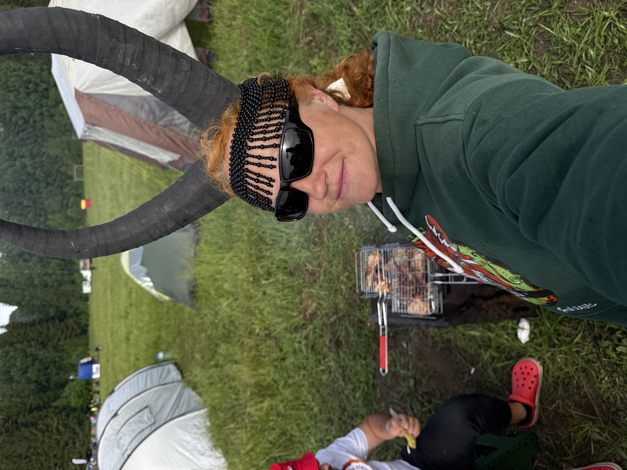This image captures an outdoor scene at what appears to be a campsite or festival in Bol'shaya Kivara, Russia. The primary subject is a person with red hair, seen from the chest up, wearing dark sunglasses and a distinctive black beaded headpiece that frames their face. They are dressed in a dark green hoodie, which has "Good Style" visible in white text on the sleeve. Two prominent, dark, horn-like structures extend from near their head, suggesting a costume or decorative accessory. The person has a subtle, content smile.
In the background, a grassy field is dotted with several tents of various sizes, indicating a camping setup. Beyond the tents, dense green trees line the horizon. To the right of the main subject, a portable barbecue grill is in use, with food (likely meat pieces) cooking on its grate. Partially visible in the lower right corner is another individual wearing black pants and red Crocs, seemingly engaged with the grill or holding an item nearby. The ground in the foreground appears damp and muddy in spots, and the lighting suggests an overcast daytime environment. The overall atmosphere is casual and indicative of an outdoor gathering.