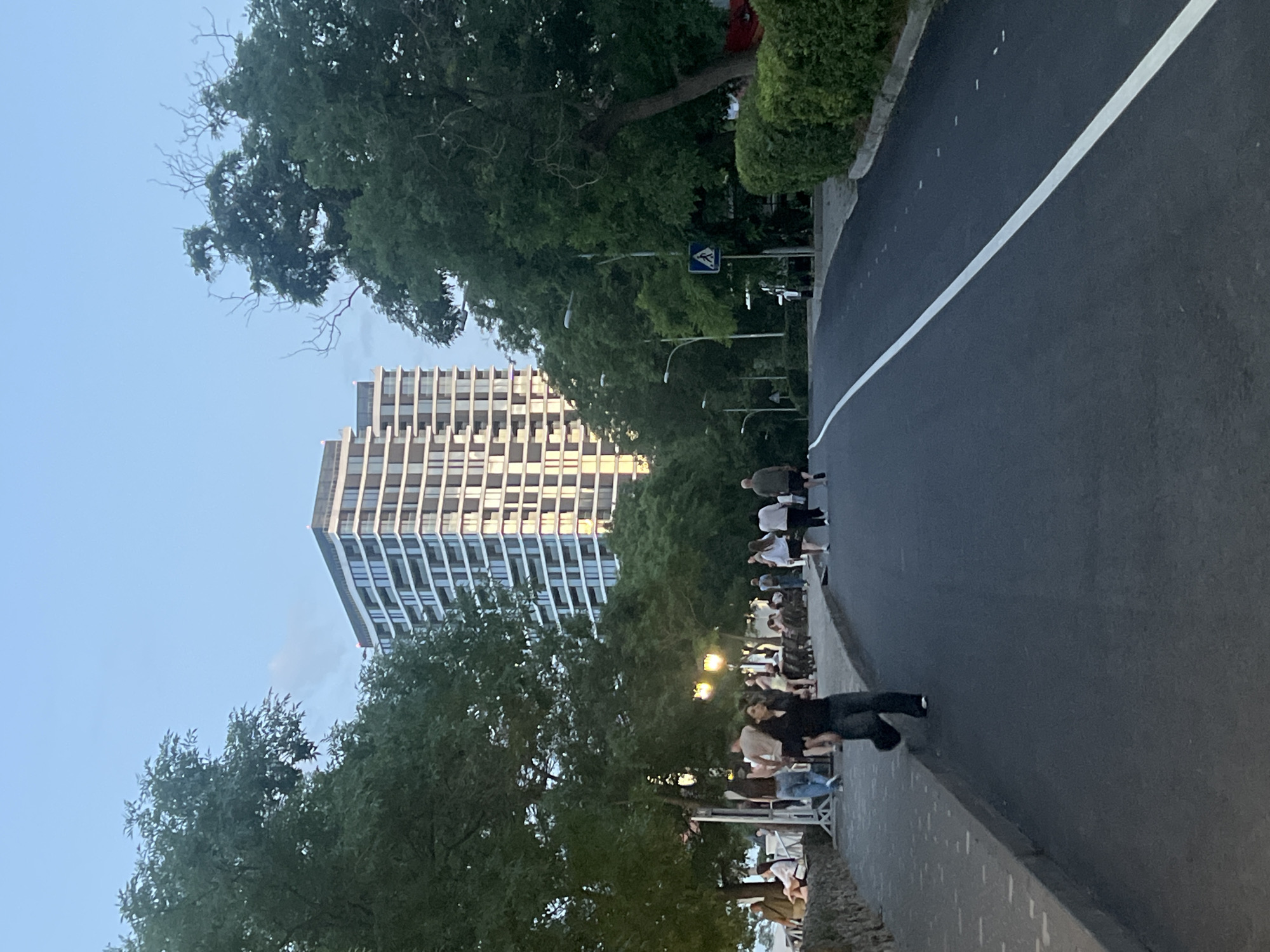 The image depicts an urban scene in Odesa, Ukraine, possibly during the late afternoon or early evening. A modern, multi-story building with a distinctive facade of horizontal bands of windows and balconies dominates the background. The building is flanked by lush green trees. In the foreground, a paved road with a white dividing line runs through the scene. To the left of the road is a raised pedestrian walkway with a stone retaining wall, where several people are walking and sitting. A woman in a dark top and dark pants is prominently featured walking on the pedestrian path. Further down the path, a group of people are casually strolling. On the right side of the road, a well-maintained hedge lines the street, and a pedestrian crossing sign is visible. Streetlights are also present along the walkway. The sky is clear with a few wispy clouds, suggesting fair weather.