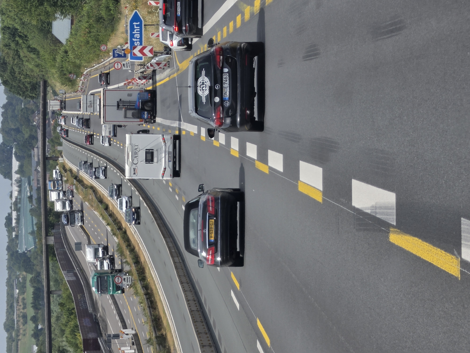The image shows a multi-lane highway with traffic in Königswinter, Germany. In the foreground, a black convertible with license plate R-539-GJ is driving on the right lane. To its right, a black station wagon with license plate DN-E 7463 is in the next lane. In the background, several trucks, cars, and a recreational vehicle are visible. The highway is surrounded by greenery and trees, with an overpass visible in the distance. Road signs indicating speed limits and lane closures are present. The sky is overcast.