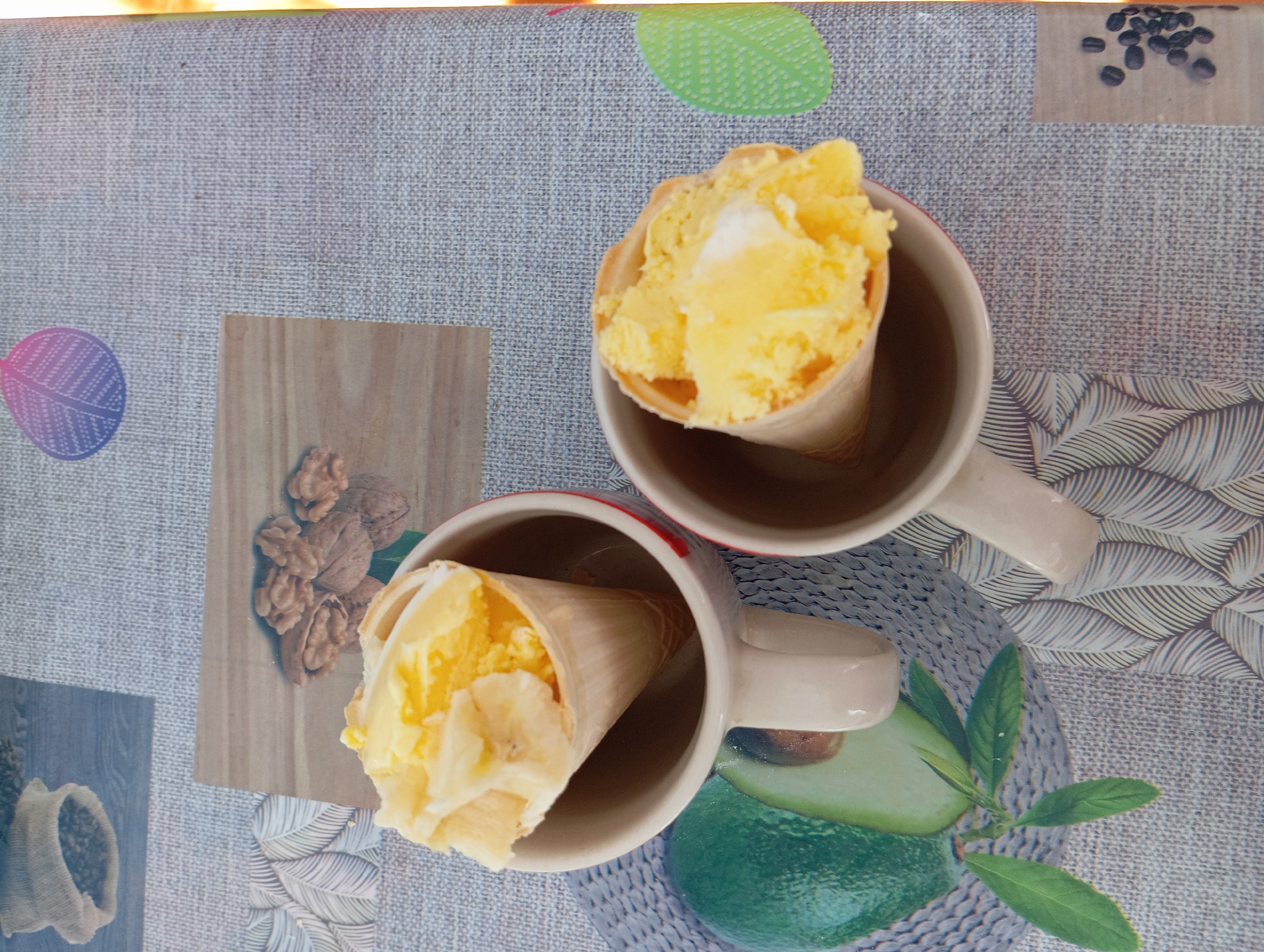 The image displays two ice cream cones filled with yellow ice cream and banana pieces, placed inside two mugs on a table. The table surface features a patterned cloth with various decorative elements, including images of walnuts, an avocado, and coffee beans. The scene appears to be set outdoors on a sunny day, as indicated by the lighting and the presence of a leaf-like decoration on the table. The specific location within the city of Chortkiv, Ukraine, cannot be determined from the image alone.