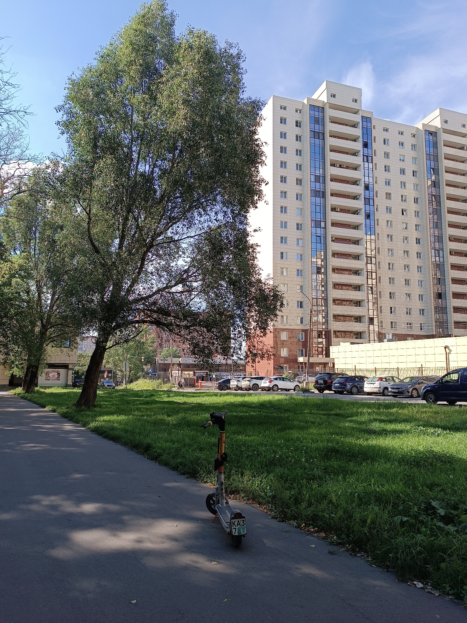 The image depicts a residential area in Zhukovsky, Russia, on a sunny day. A large, multi-story apartment building with a beige facade and blue-tinted windows dominates the background. In the foreground, a paved walkway runs alongside a grassy area where several cars are parked. A green electric scooter with a license plate is visible on the walkway, casting a shadow. Large trees with dense foliage are scattered throughout the scene, providing shade. The sky is clear blue with a few wispy clouds.