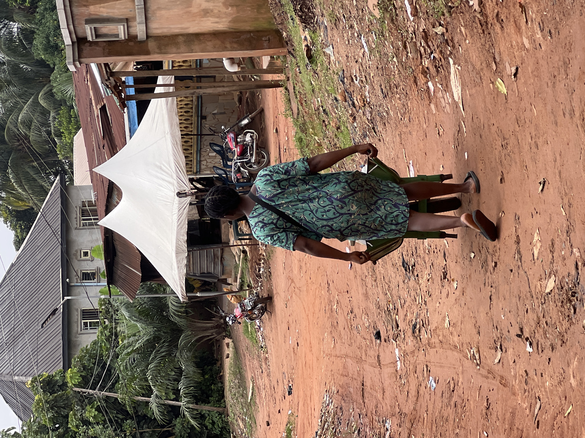 In Nkpor, Nigeria, a woman walks away from the camera on a dirt path, pushing a green wheelbarrow. She wears a green and purple patterned dress and dark flip-flops. The path is littered with debris, including leaves, plastic, and discarded items. In the background, a white tent stands over a structure with a corrugated metal roof. A red motorcycle is parked near the tent, and beyond that, buildings with grey and brown walls are visible, some with corrugated roofs. Lush green foliage, including palm trees, surrounds the area. The sky is overcast.