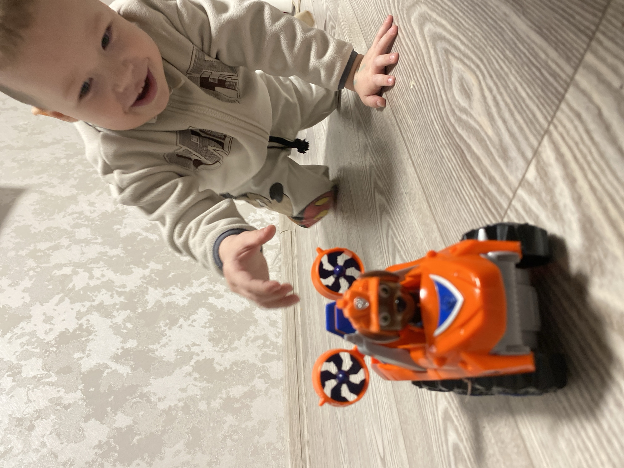 A young child wearing a light-colored zip-up sweatshirt and pants is reaching towards an orange toy vehicle. The toy has propellers and black wheels. The floor is a mix of light wood-grain and speckled white and beige patterns. There is no indication of Sochi, Russia.
