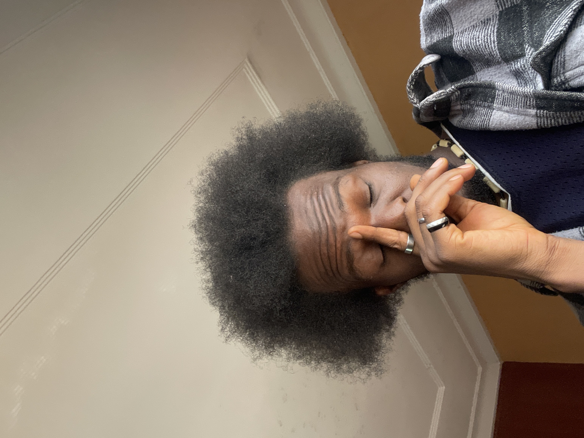 The image shows a close-up, medium shot of a Black man with a large afro hairstyle. He is positioned in the foreground, with his face tilted downwards and his eyes closed. His right hand is raised to his face, with his fingers touching his forehead and nose, suggesting he might be feeling tired or unwell. He is wearing a dark blue undershirt and a grey and black plaid overshirt, with a necklace made of light-colored beads visible. The background features a textured ceiling with decorative molding and a portion of a wall painted in a warm, orange hue. The lighting suggests an indoor setting, possibly during daylight. The image does not provide any specific visual cues to pinpoint its location within Jos, Nigeria.