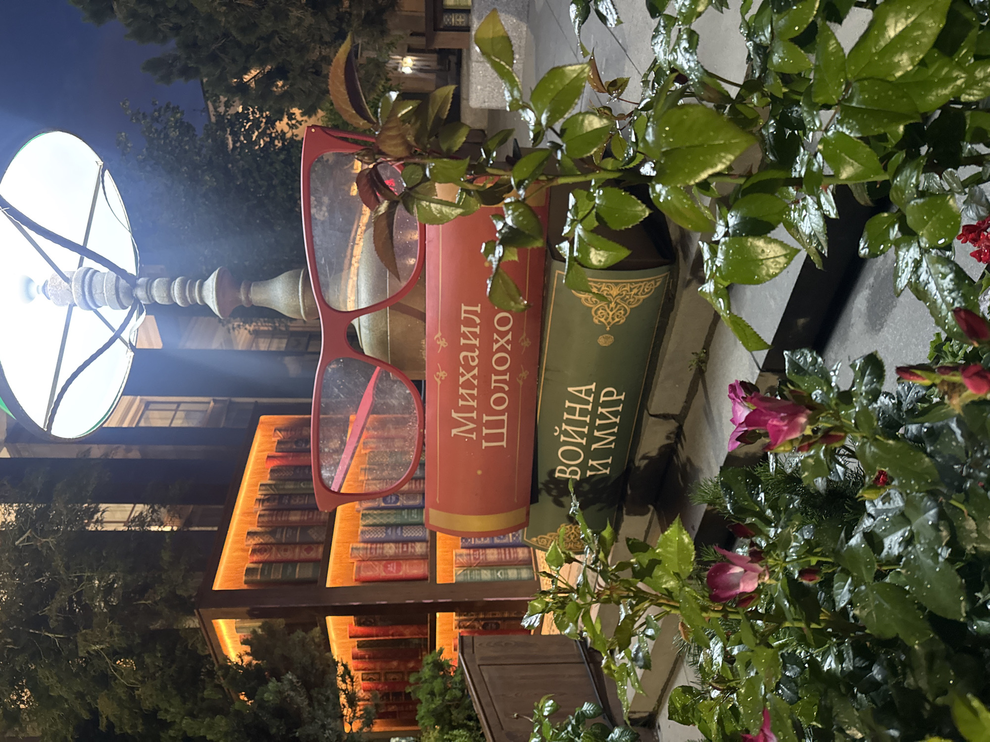 The image depicts an outdoor display featuring oversized books and red-rimmed eyeglasses, set against a backdrop of illuminated bookshelves. The books are stacked vertically, with the top book in red inscribed with Russian text that appears to be "Михаил Шолохов," and the book below it in green with the Russian text "ВОЙНА И МИР." The eyeglasses are placed atop the stack of books. Behind the display, a lamp post with a large white lampshade is visible. The illuminated bookshelves in the background are filled with books and appear to be part of a decorative facade. In the foreground, rose bushes with pink blossoms and green leaves, some with water droplets, are present. The scene is illuminated by artificial lighting, creating a nocturnal ambiance. The location is Moscow, Russia, specifically the "Bulgakov House" museum onogradny Boulevard.