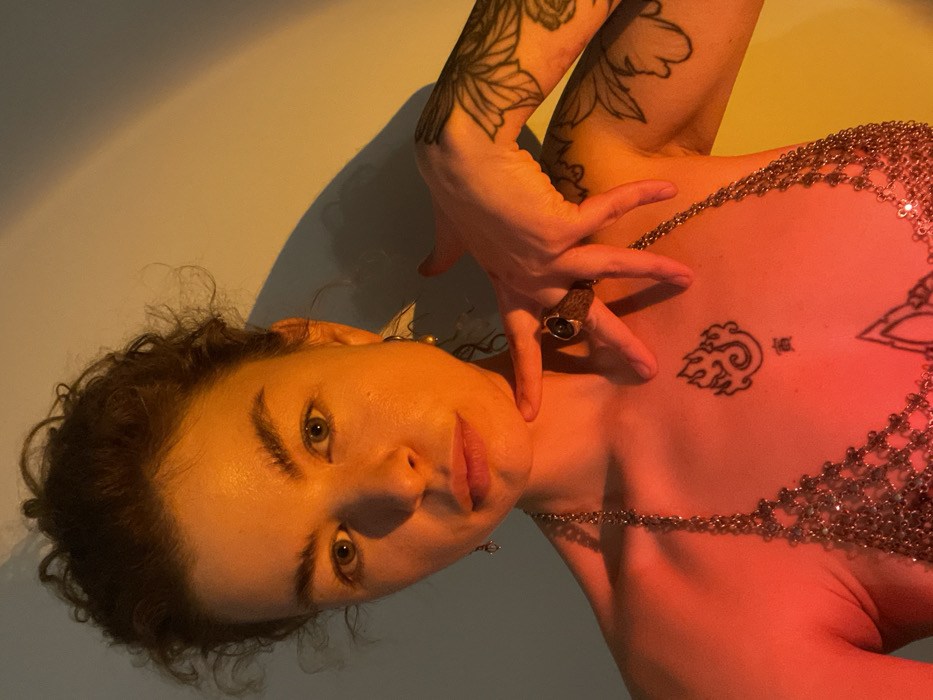 The image displays a close-up portrait of a female individual against a plain, artificially lit background. The subject has fair skin, curly dark hair pulled back, and light-colored eyes, gazing directly forward. She wears a metallic, possibly chainmail, halter-style top. The right side of her body, including her left shoulder and chest area, is illuminated by a strong red light, while the left side of her body and the background to her right are illuminated by an orange-yellow light, creating a distinct color gradient.
Jewelry includes a small drop earring on her right earlobe and a larger, abstract, possibly geometric or triangular earring with a small bead on her left ear. Her right hand is raised, with the index finger touching her chin/jawline. A prominent, dark-colored ring is worn on the index finger of her right hand. Visible tattoos include a stylized flame or abstract symbol with a small character underneath on her sternum, an outline floral or organic sleeve design on her left arm, and a partial outline tattoo below the sternum. Shadows are cast on the wall behind the subject, indicating directional lighting. The environment is an indoor setting with controlled lighting.