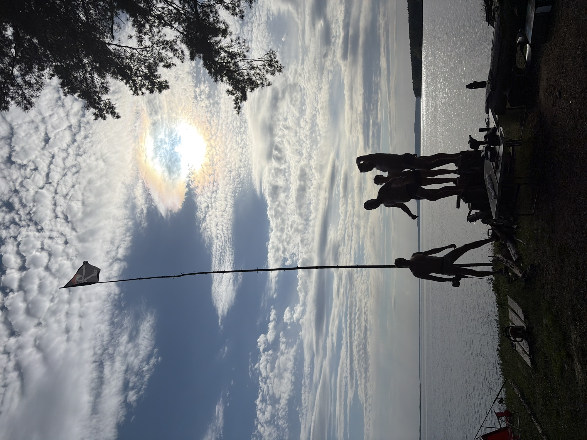 The image shows a lakeside scene under a partly cloudy sky with a solar halo visible around the sun. Silhouetted figures of people stand on a structure near the water's edge, possibly a dock or platform. A long pole extends into the sky with a flag bearing a skull and crossbones design. Trees are visible in the upper left corner and along the shoreline. Based on the request, the location is Zabegaevo, Russia.