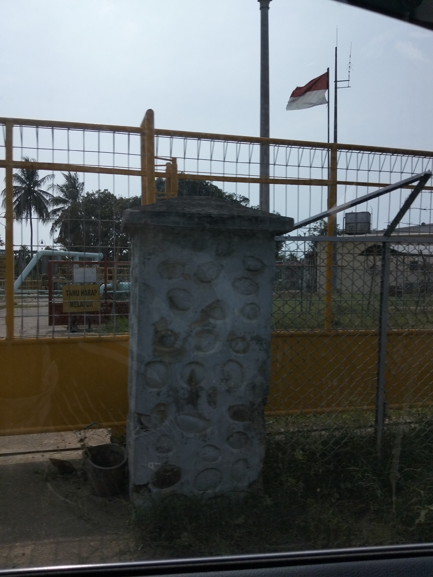 This image depicts an outdoor scene in Blang Mee, Indonesia, focused on a gate and surrounding industrial or utility area. A large, yellow fence with a diamond mesh pattern encloses the property. In the foreground, a weathered concrete pillar, textured with circular indentations, stands to the right. Behind the pillar and fence, a red gate is visible with a yellow sign that reads "TAMU HARAP MELAPOR," meaning "Visitors Please Report." Beyond the gate, pipes and industrial equipment are present, suggesting a functional facility. A red and white Indonesian flag flies on a pole in the background, indicating national presence. Coconut palm trees are visible on the left, typical of the region. The sky is overcast. The scene appears quiet and not actively populated.