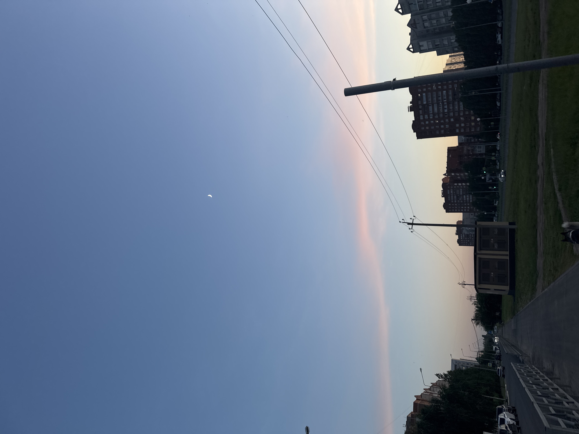 The image captures a cityscape at dusk. The sky transitions from a deep blue to a pale orange-pink, with a crescent moon visible in the blue section. Electrical wires stretch across the sky. On the right, buildings of varying heights line the street, with some trees interspersed. A paved road and a grassy area run parallel to the buildings. A pole is visible in the foreground on the right side of the frame. There appear to be cars parked along the street and some are moving. The image is shot from ground level.