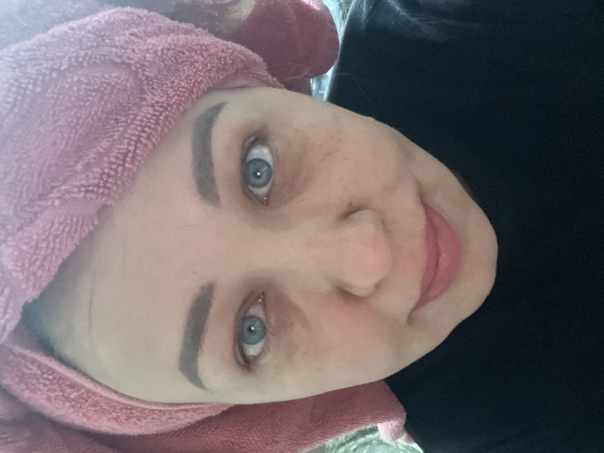 A person with light skin and blue eyes is facing the camera, their head wrapped in a pink towel. The person has freckles and defined eyebrows. They are wearing a black top. The background is indistinct. There is no contextual information to suggest the location is Chaikovsky, Russia.