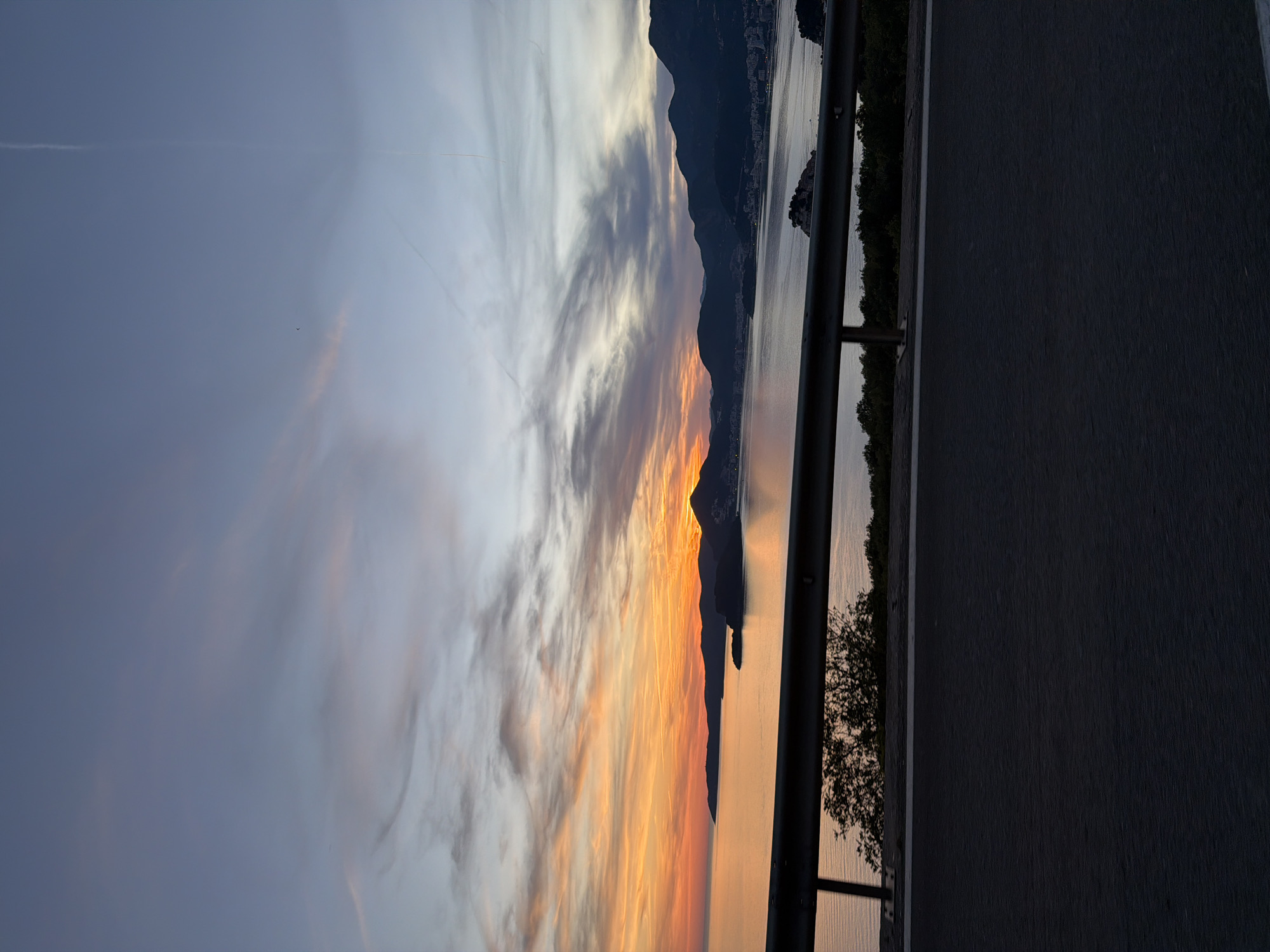 The image captures a scenic view of a coastal landscape at sunset, likely in the vicinity of Blizikuće, Montenegro. The foreground features a dark asphalt road and a metallic guardrail, suggesting the perspective is from a road overlooking the sea. Beyond the guardrail, a body of calm water, likely the Adriatic Sea, stretches towards the horizon. The water reflects the warm hues of the setting sun.
A rugged coastline with dark, silhouetted mountains forms the backdrop. Nestled along the coast is a town or settlement, with buildings faintly visible against the darkening landscape. A small island or rocky outcrop is present in the water, adding to the picturesque setting.
The sky above is a dramatic canvas of color. The left side of the sky is dominated by vibrant orange and yellow streaks, indicative of the setting sun. These warm colors gradually transition to softer pinks and purples, and then to a muted blue-grey in the upper portion of the sky. Wispy clouds are scattered across the sky, catching the last rays of sunlight. A faint contrail is visible in the upper right section of the sky.
The scene is devoid of people or any discernible action, presenting a static, natural landscape illuminated by the twilight. The overall impression is one of tranquility and natural beauty during the transitional period between day and night.